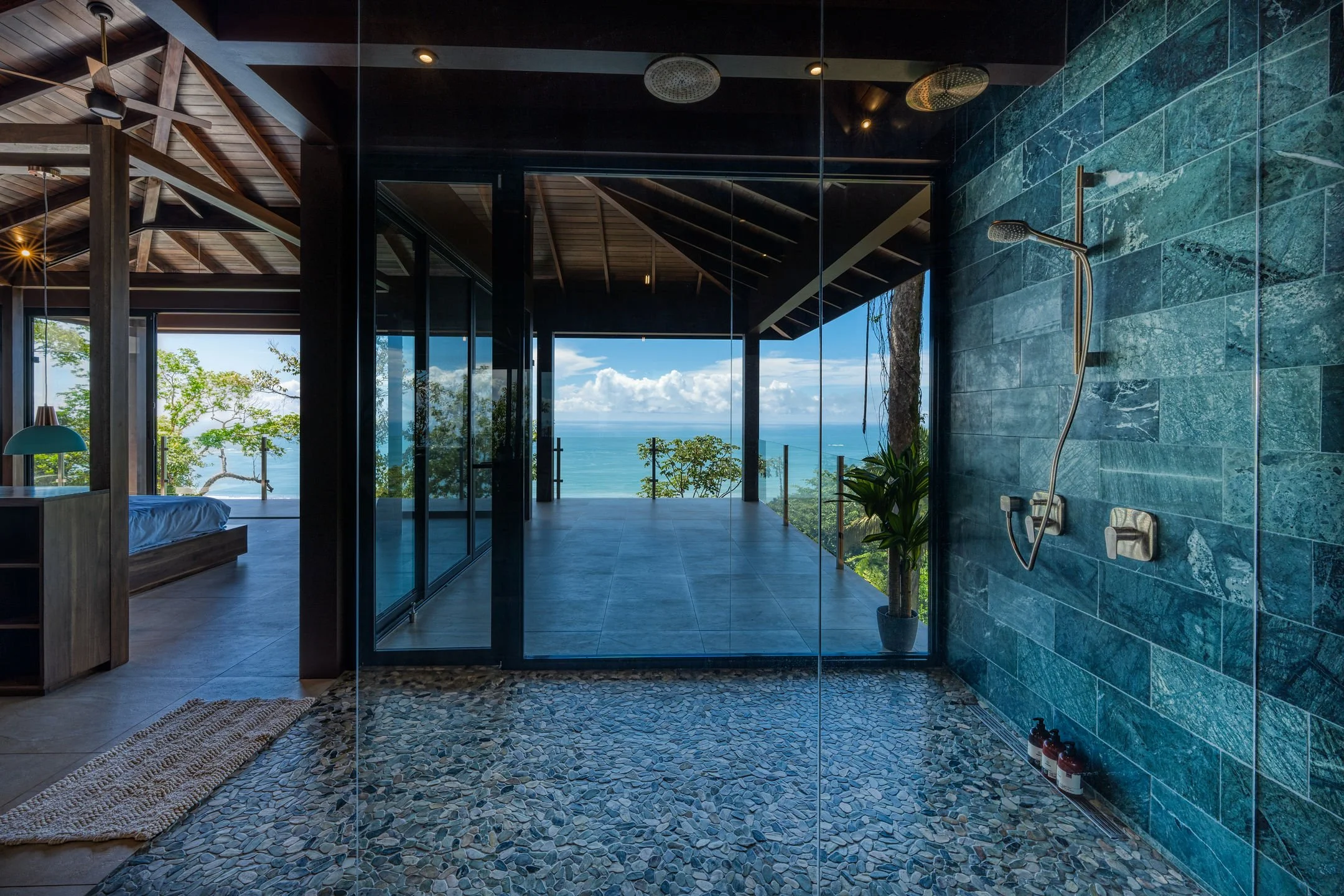 Open-air shower with blue-green tiled wall, pebble floor, and ocean view on a balcony