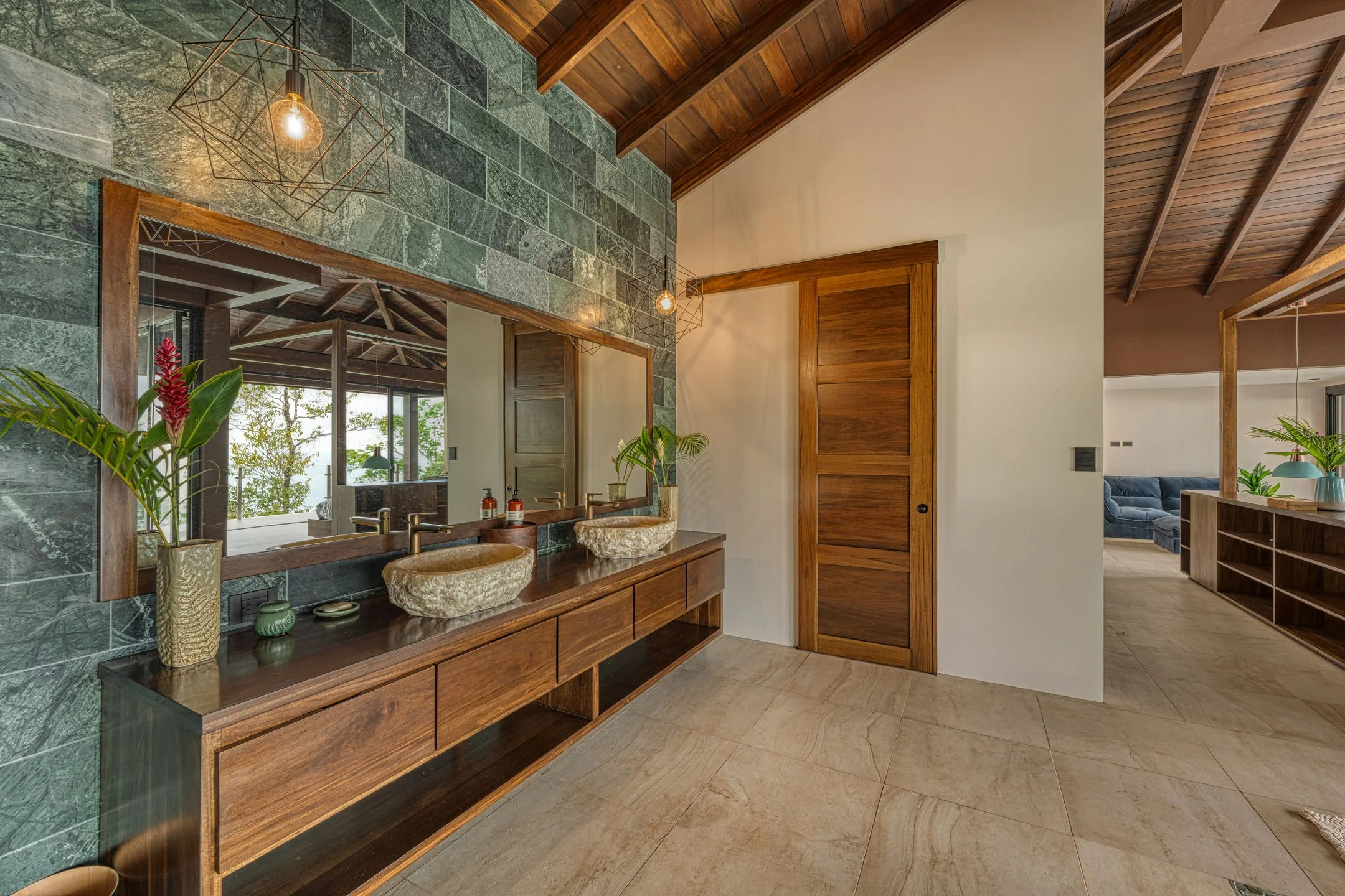 A bathroom with a large wooden vanity, two stone vessel sinks, and a wide mirror. There are potted plants and soap dispensers on the vanity. The wall behind the mirror has green stone tiles, and there is a view of a living area with large windows and a blue sofa in the background. The ceiling is wood-paneled.