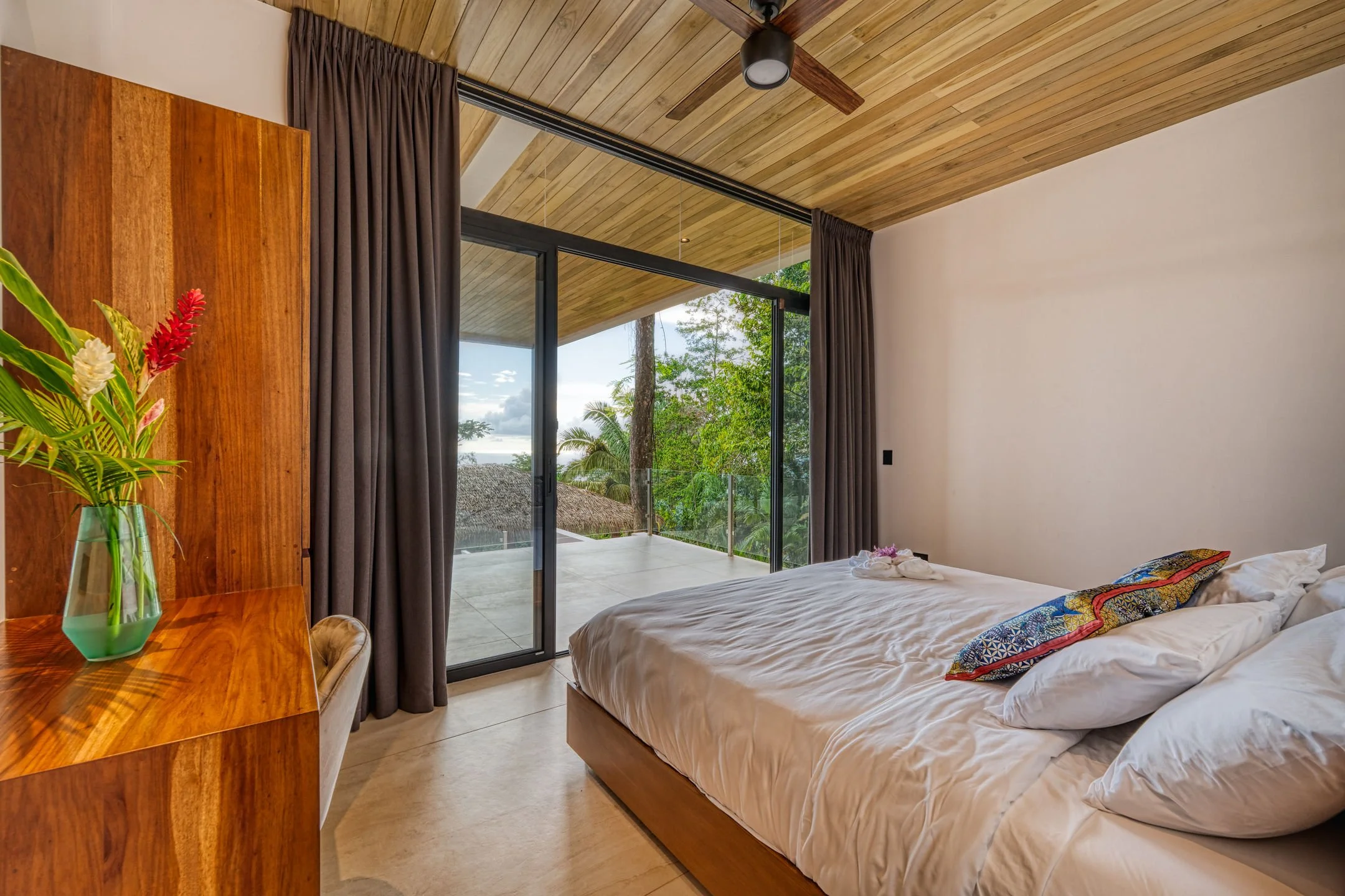 A bedroom with a wooden ceiling, a large sliding glass door leading to a balcony, a white bed with colorful pillows, a bedside towel decoration, a wooden dresser with a glass vase of tropical flowers, and dark curtains.