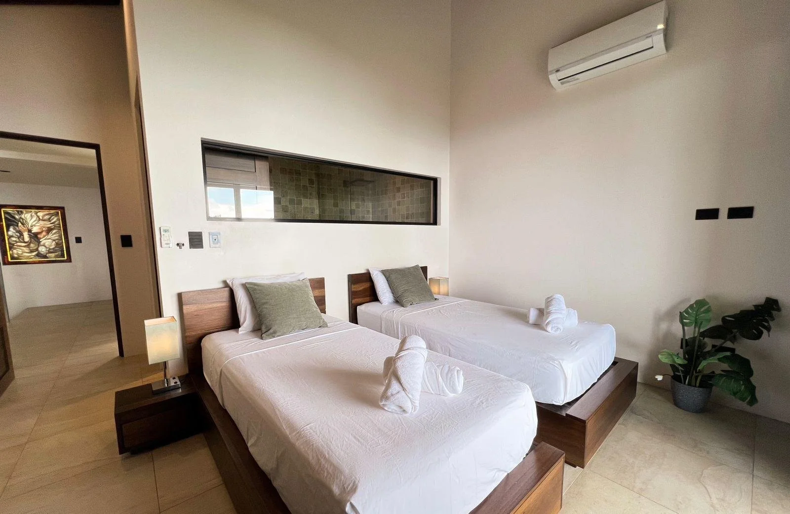 A modern hotel room with two single beds with wooden headboards, white linens, gray pillows, and rolled towels on each bed. There are nightstands with lamps, a large window behind the beds, and a potted plant in the corner. An air conditioning unit is mounted on the wall.