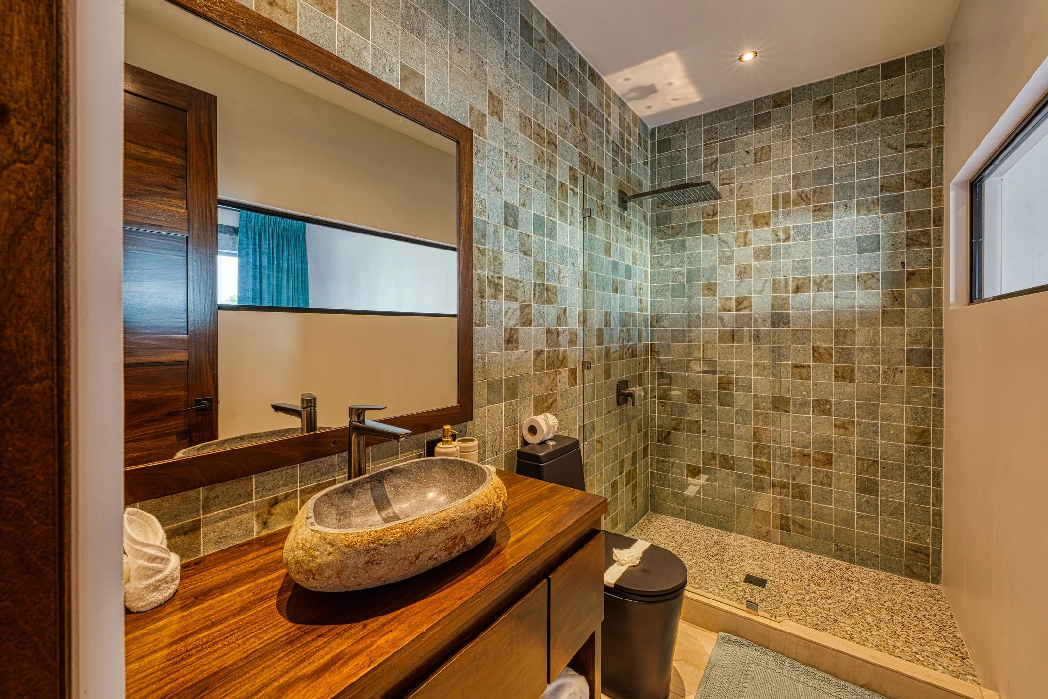 Modern bathroom with a wooden vanity, stone vessel sink, large mirror, and walk-in shower with green tile walls and a window.