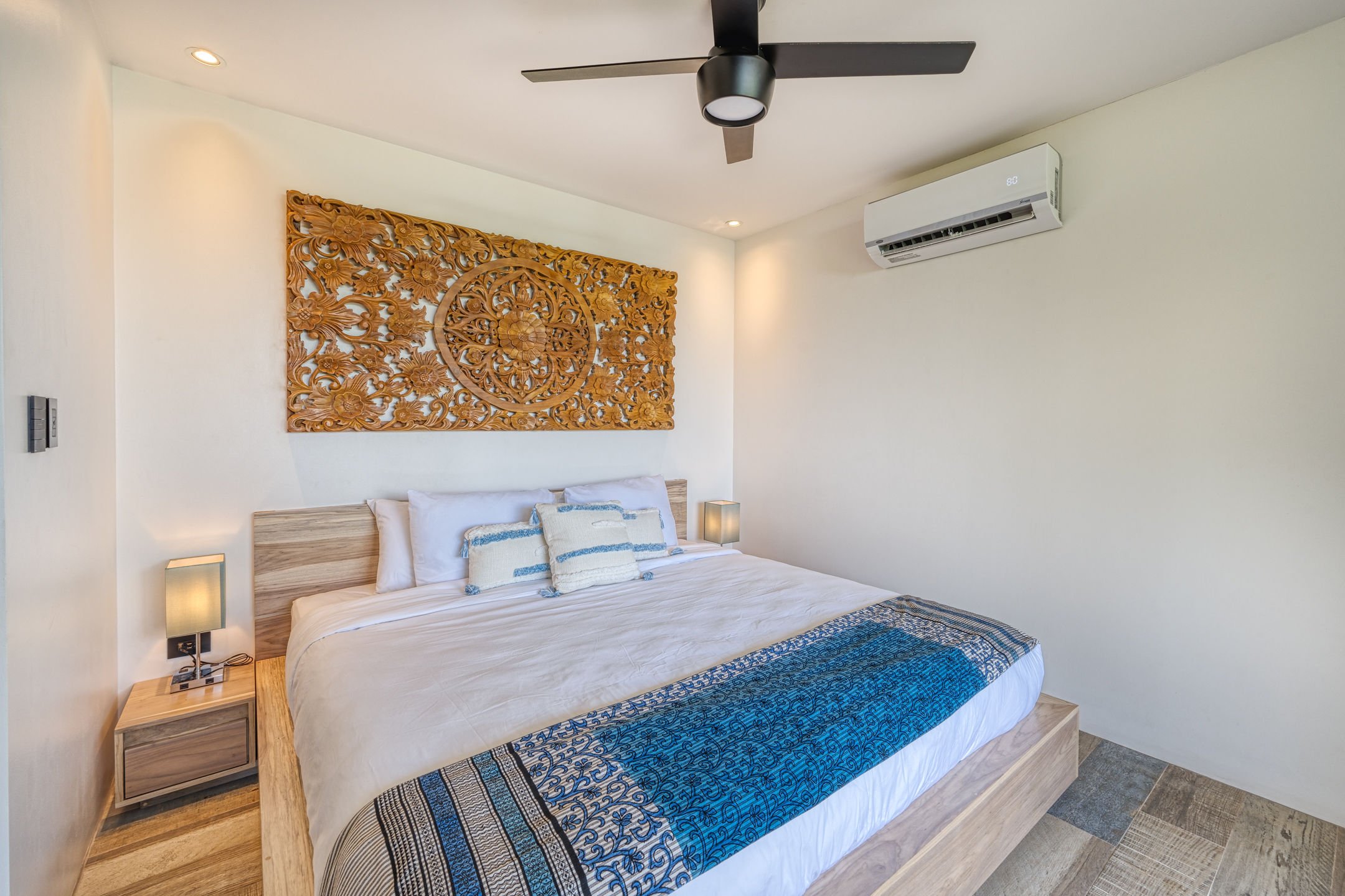 A bedroom with a large bed featuring white bedding and decorative pillows, wooden headboard, two bedside lamps, a wall-mounted air conditioner, a ceiling fan, and a carved wooden wall art piece above the bed.