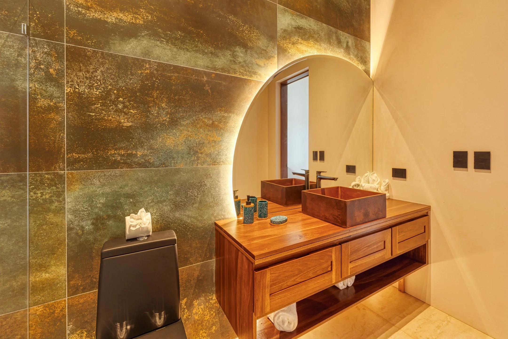 Modern bathroom with antique copper sinks, wooden vanity, large mirror, green textured wall tiles, black tissue box on stool, and decorative items on the counter.