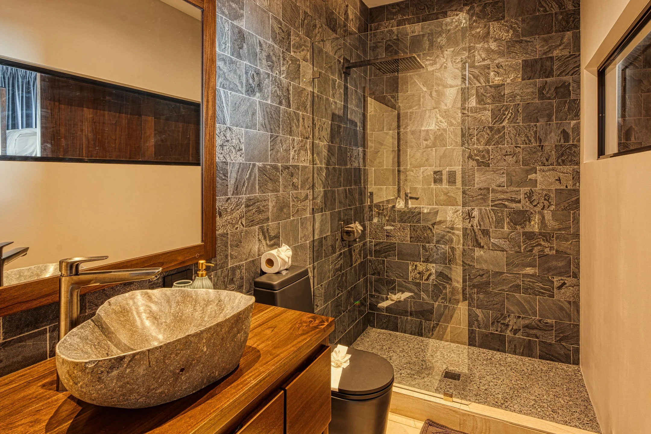 Modern bathroom with a stone vessel sink, wooden vanity, large mirror, black toilet, and glass-enclosed shower with gray stone tiles.