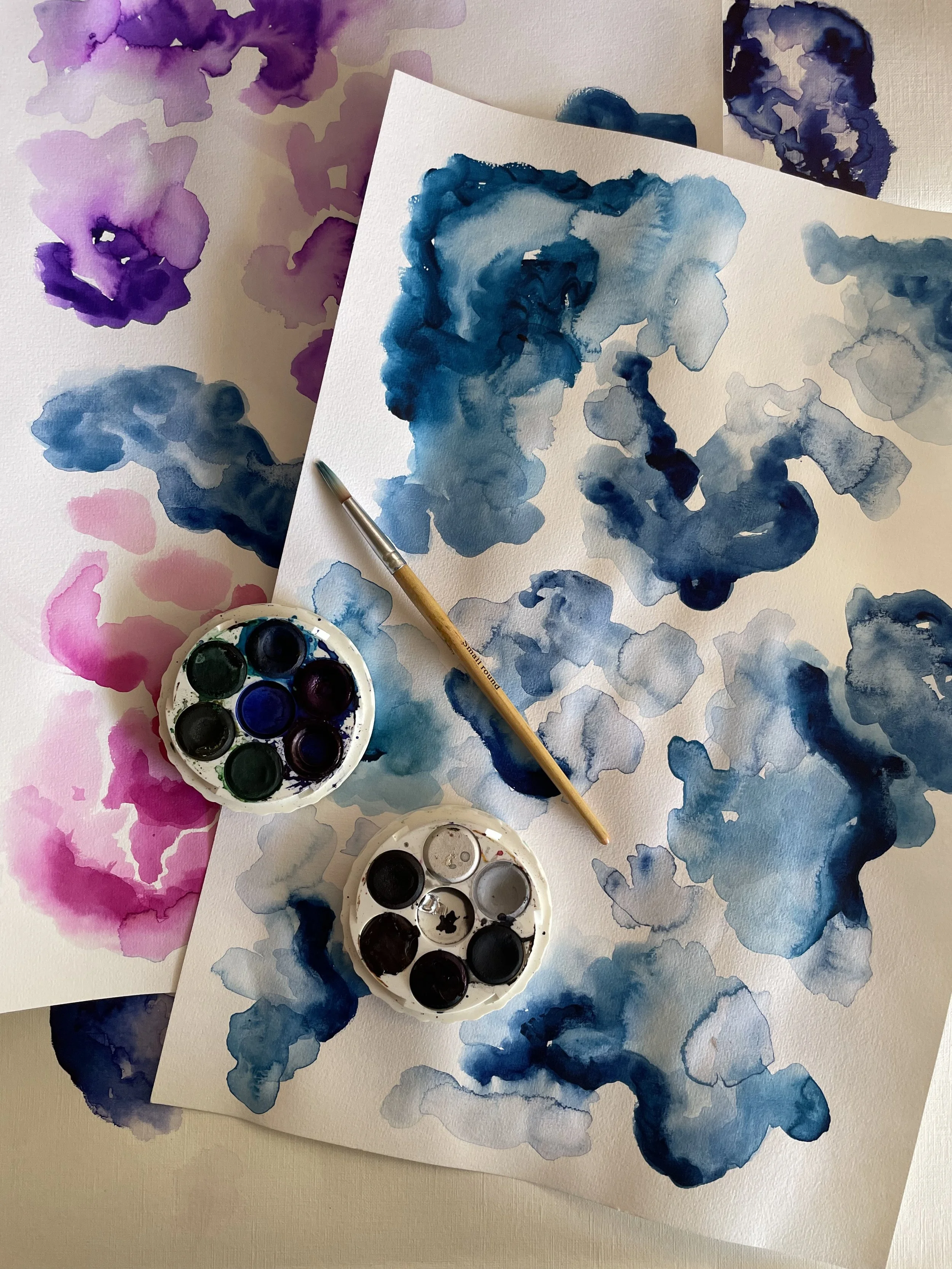 Watercolor paintings and palettes with blue, purple, and pink paint on white paper and surface.