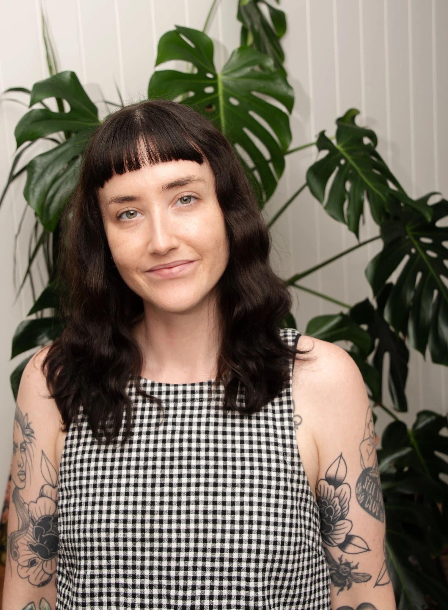 A woman with shoulder-length dark hair, light skin, and tattoos on her arms, standing in front of a large green monstera plant, wearing a sleeveless black and white checkered top.
