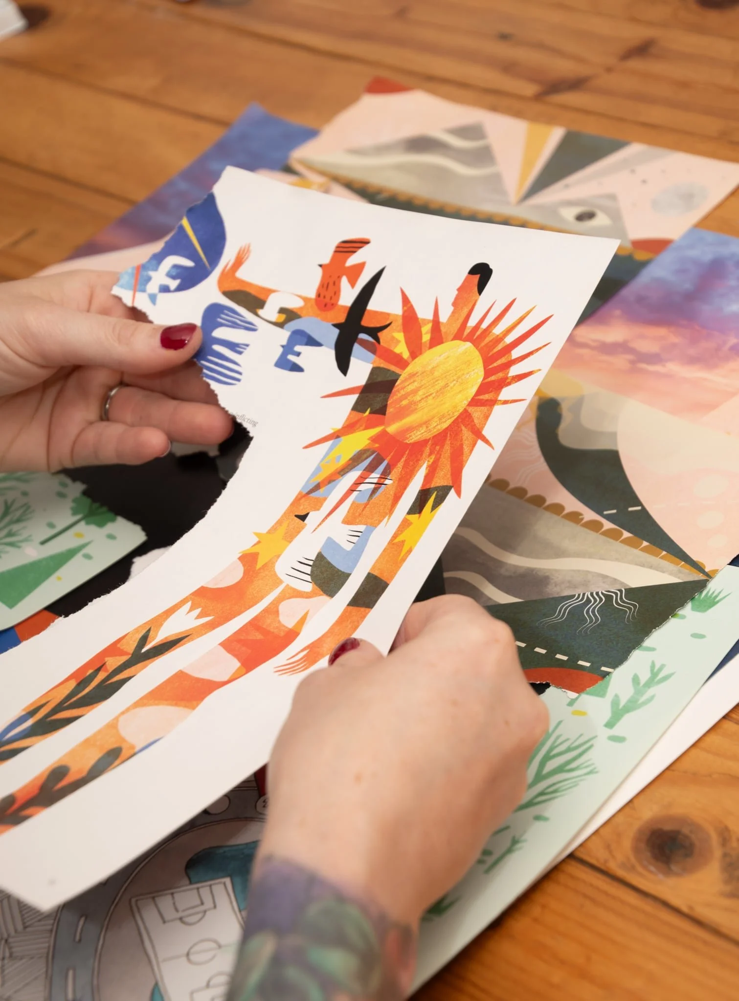 Person holding a torn colorful illustration of a person with outstretched arms, a large sun on their chest, and abstract elements, with additional colorful prints on a wooden table.