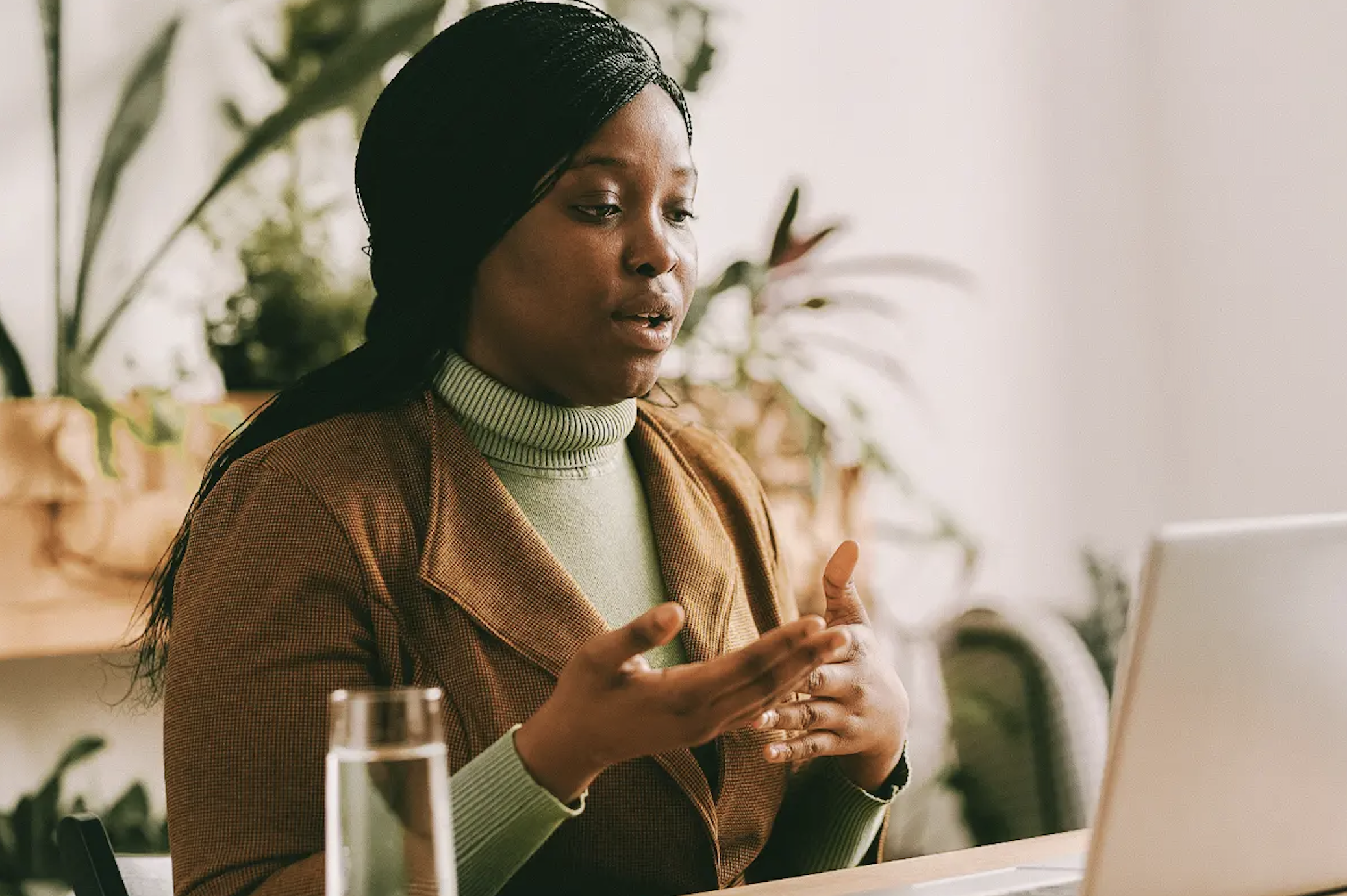 A woman with long black braided hair, wearing a green turtleneck and a brown blazer, speaking in front of a laptop in a bright room decorated with plants.