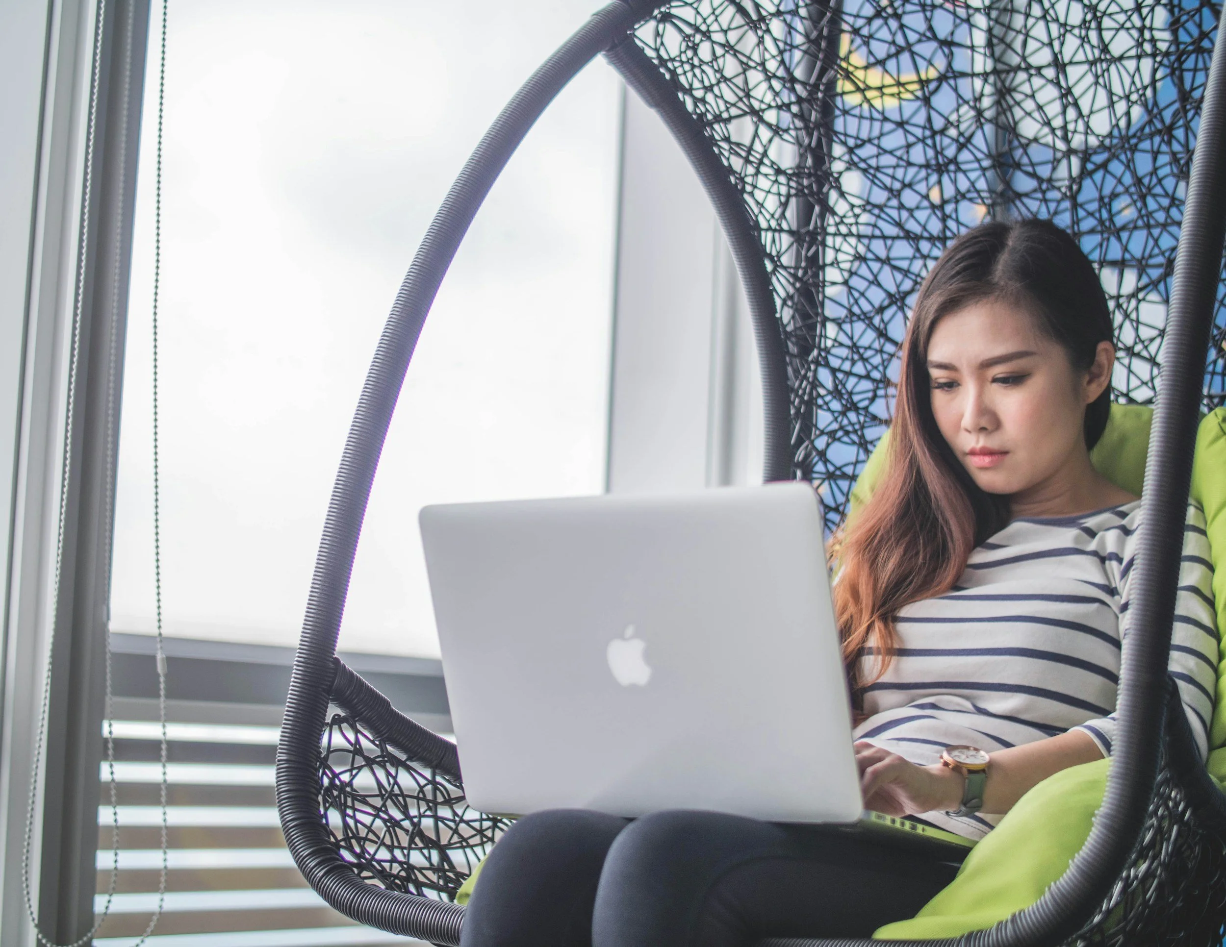 Young woman using a laptop while sitting in a hanging chair near a window.