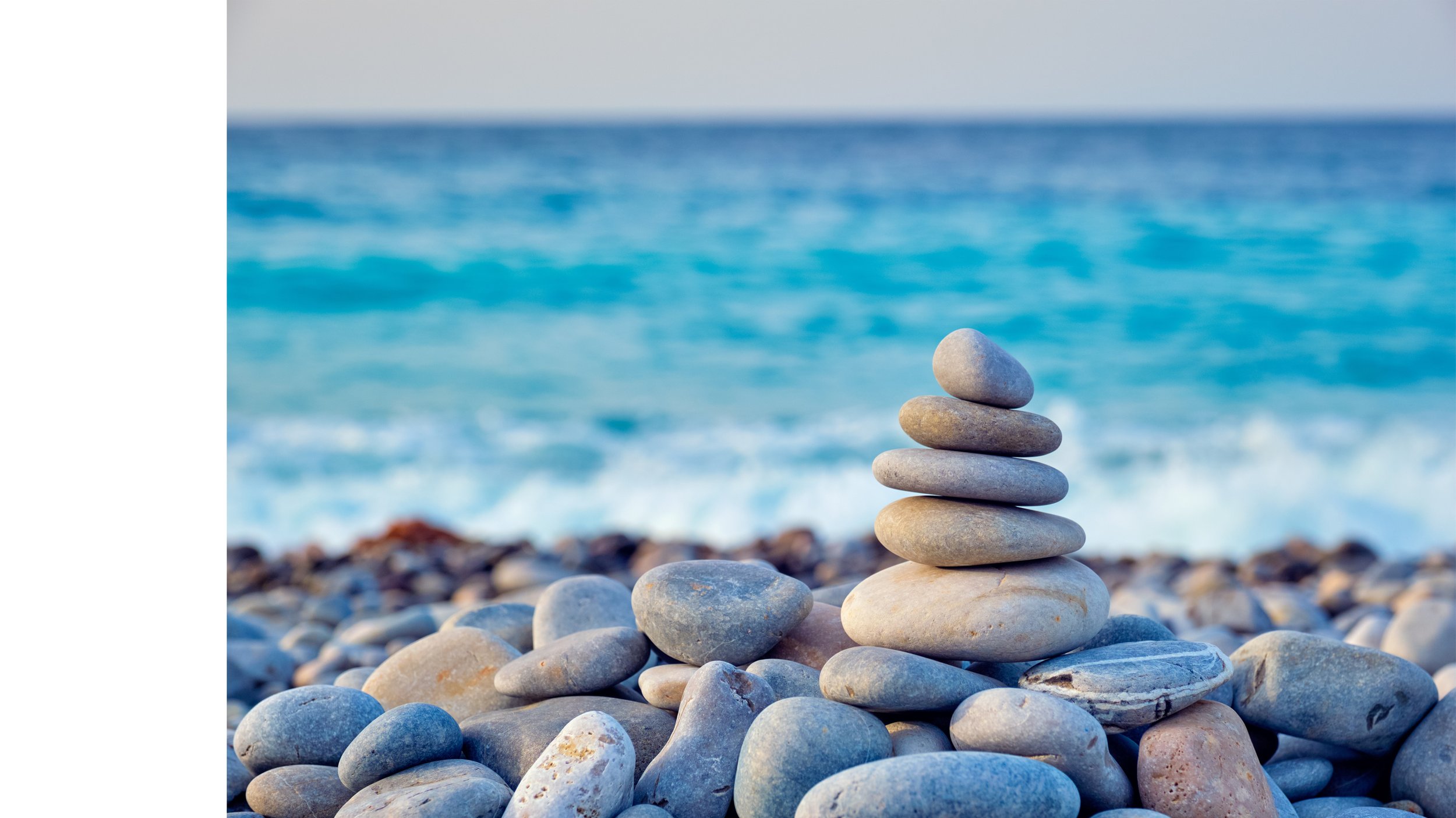 A stack of smooth, rounded pebbles balanced on a rocky beach with a blurred ocean and sky in the background.