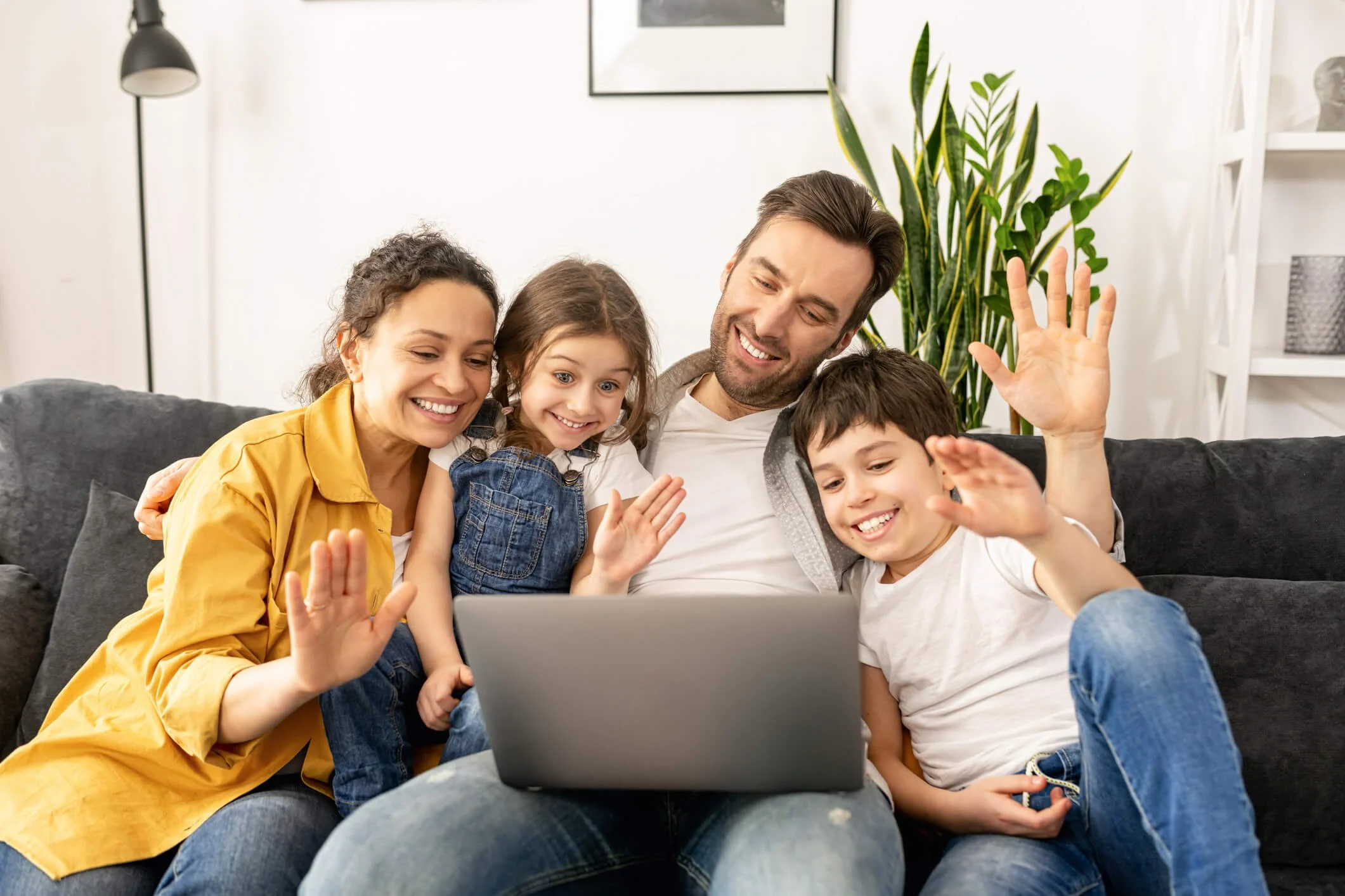 Family of four sitting on a couch, looking at a laptop, smiling, and waving, with a houseplant in the background.