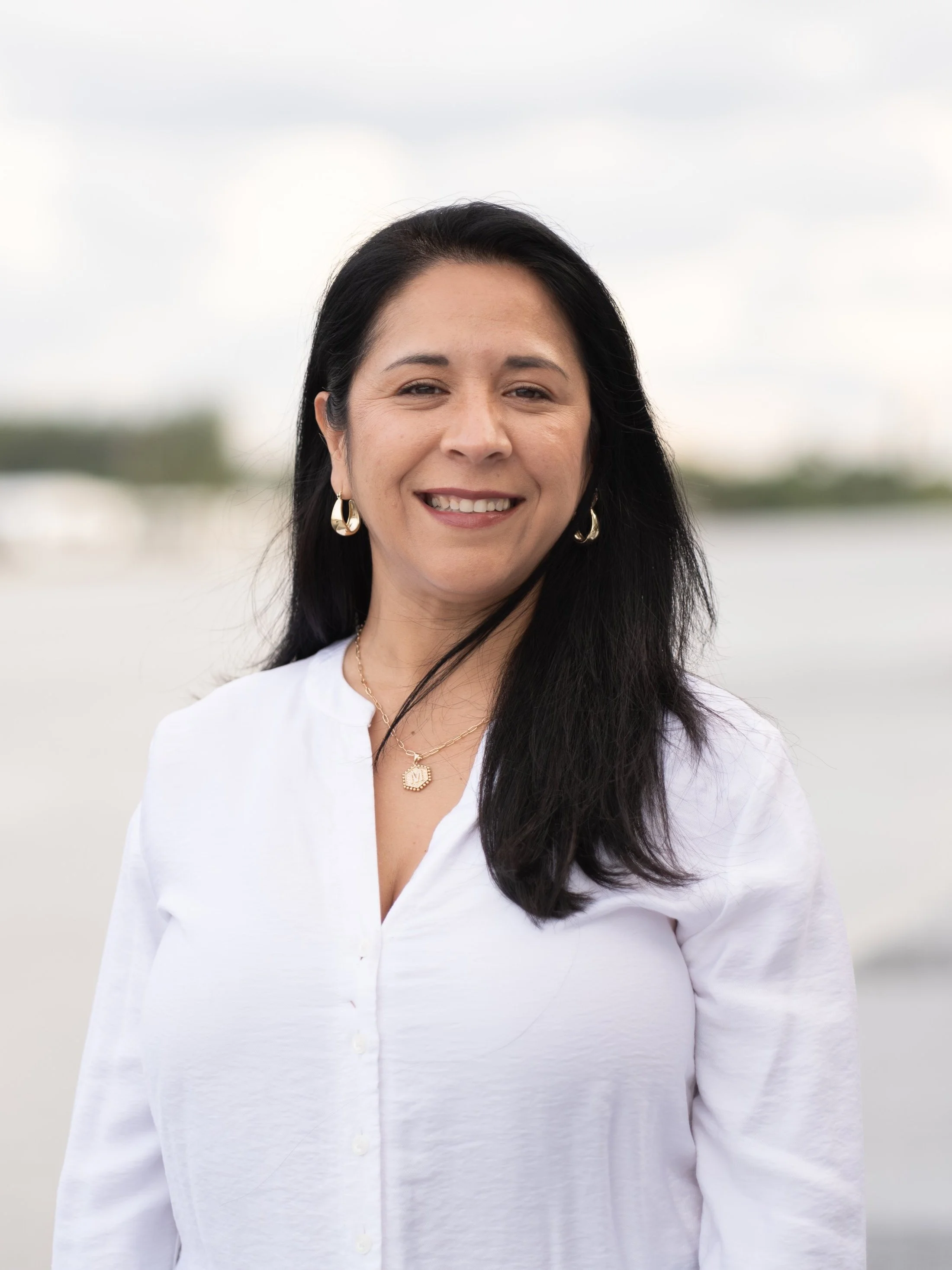 A woman with long black hair wearing a white blouse, gold jewelry, and smiling outdoors near water with a blurred background.