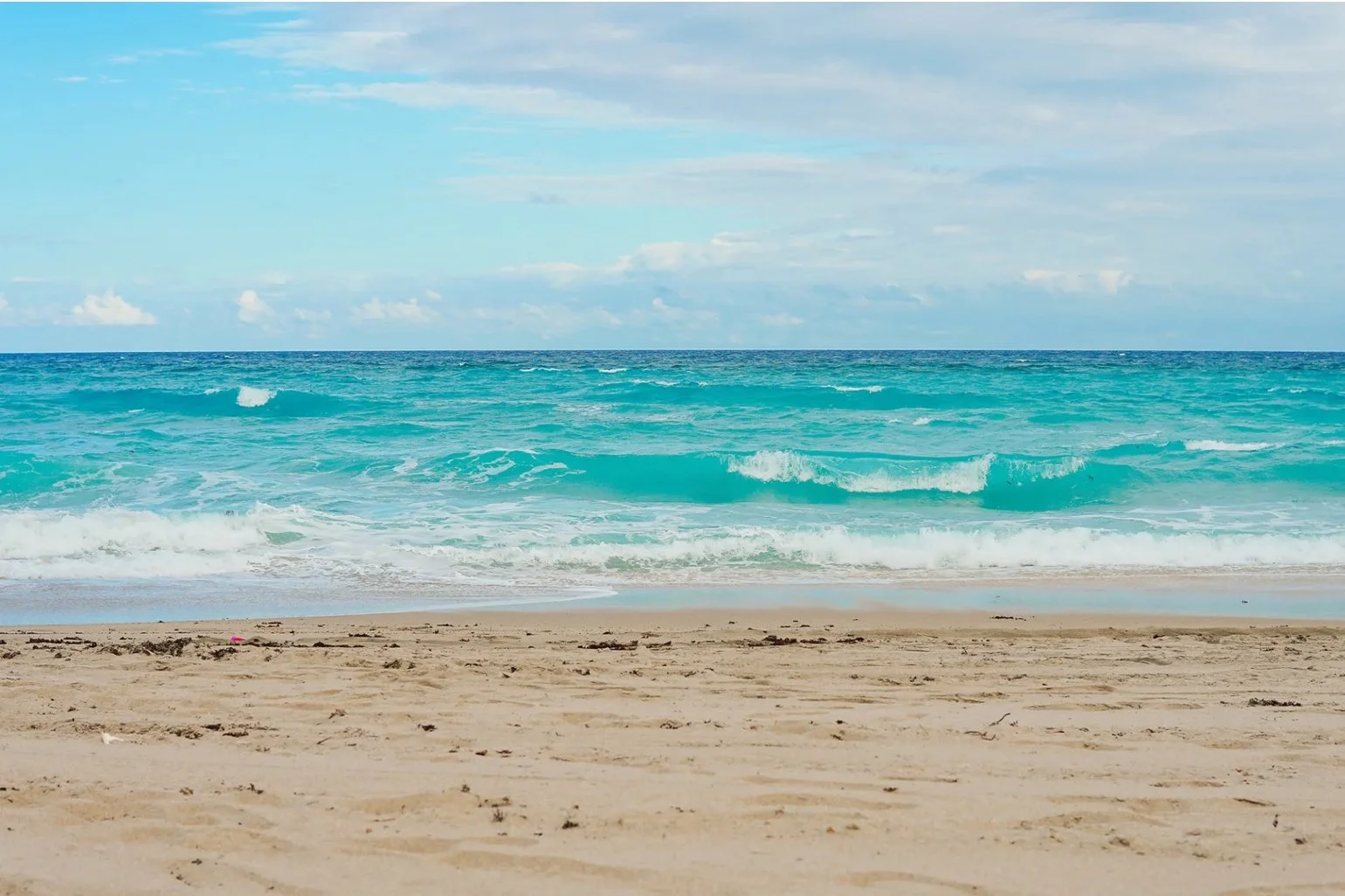 Clear ocean waves crashing onto sandy beach under partly cloudy sky.