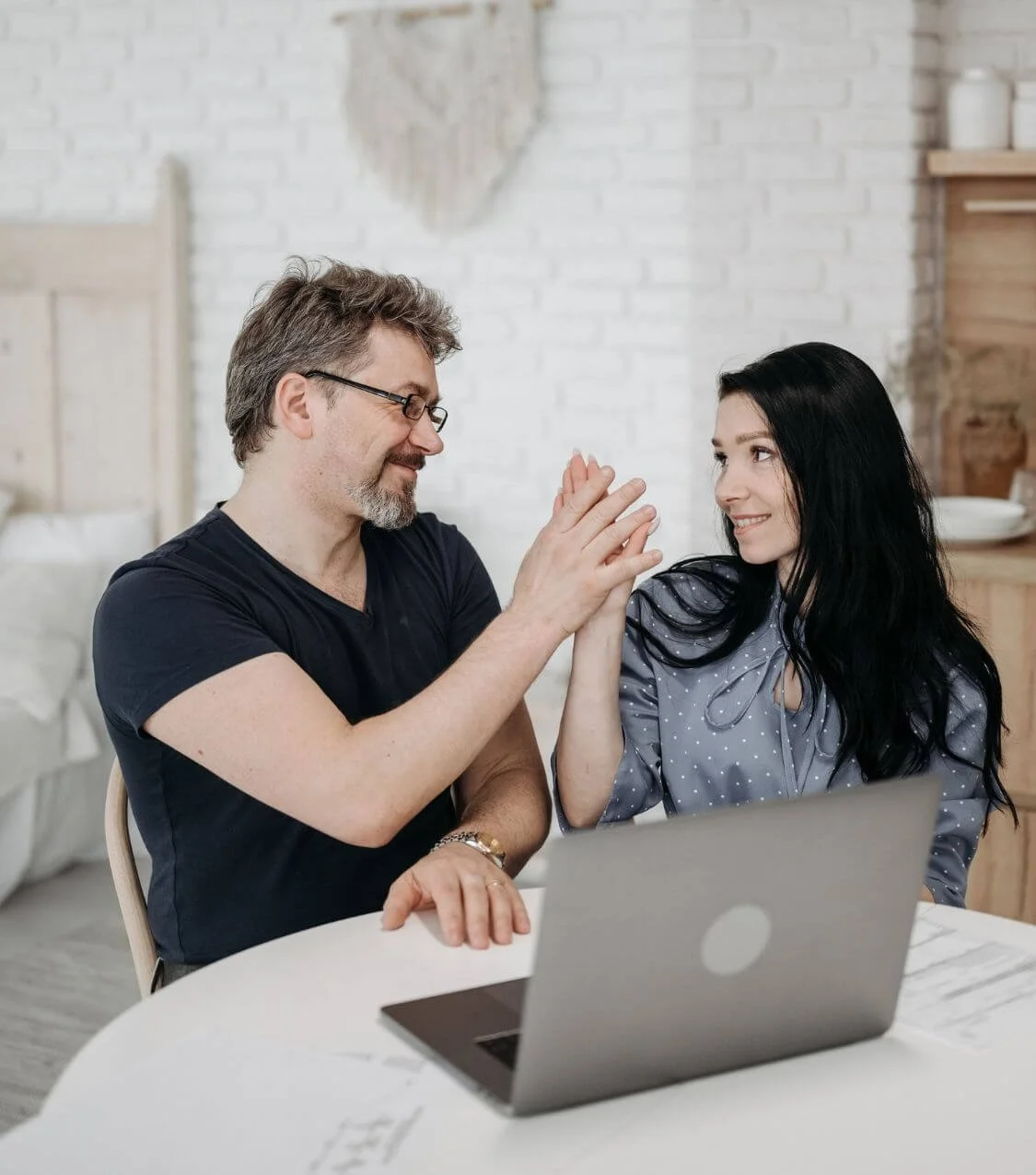 A man and woman sitting at a white table, smiling and giving each other a high five, with a laptop in front of them in a bright room with white brick walls.