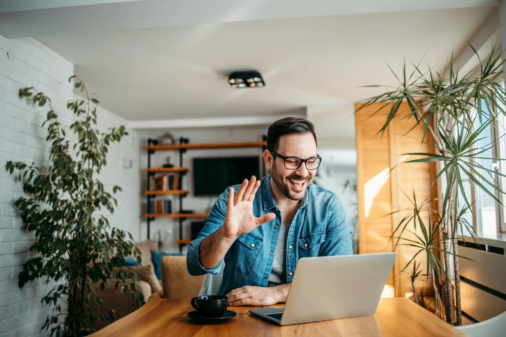 A man with glasses and a beard smiling and waving at his laptop in a bright living room with plants and a bookshelf.