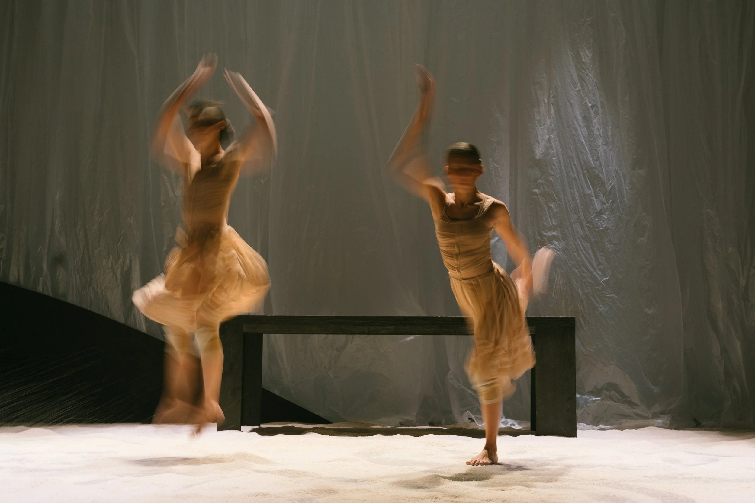 Two dancers performing on stage with a textured gray backdrop and a black bench, captured with motion blur in a contemporary dance performance.