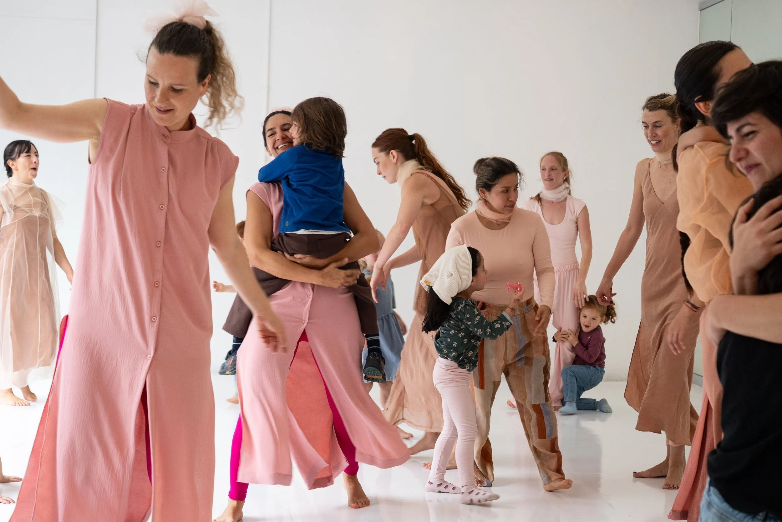 A group of women and children in a dance class, wearing flowy pastel-colored garments, some with their eyes closed and others smiling, practicing dance moves in a bright studio with a white background.