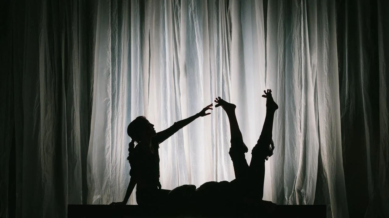 Silhouette of two children on stage, one reaching out to the other and the other lying with legs up, against a backdrop of curtains.