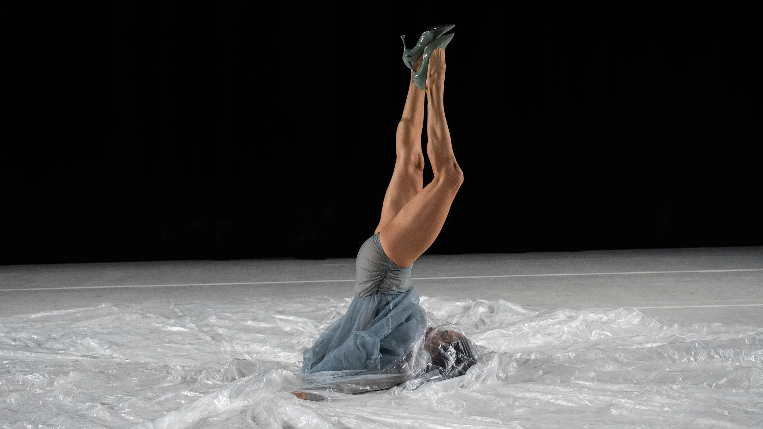 A person is lying on their back on a surface covered with plastic, with their legs extended straight up in the air. The background is black, and the person is wearing gray shorts and a gray top, with their head and arms resting on the plastic.