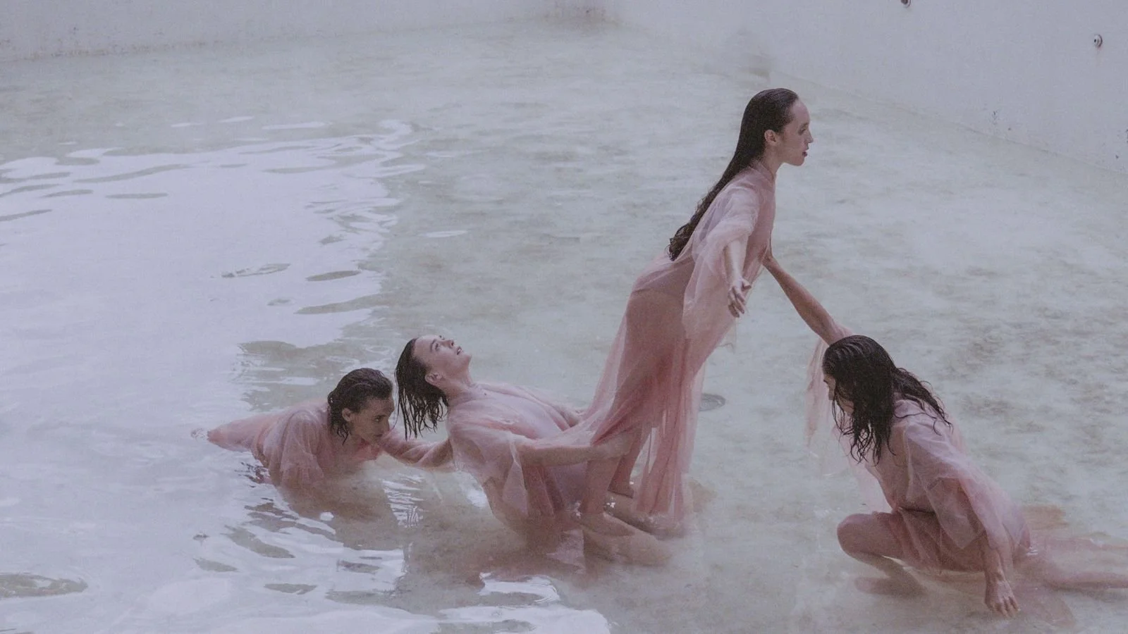 Five women wearing pink dresses are in shallow water, with some standing and some kneeling or lying down, suggesting a choreographed dance or performance.