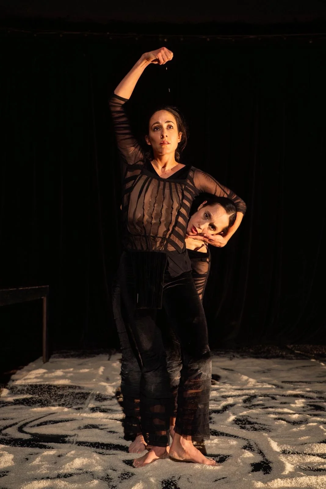 Two women dancing on a stage with white sand, wearing sheer black outfits, illuminated by stage lights, black background.