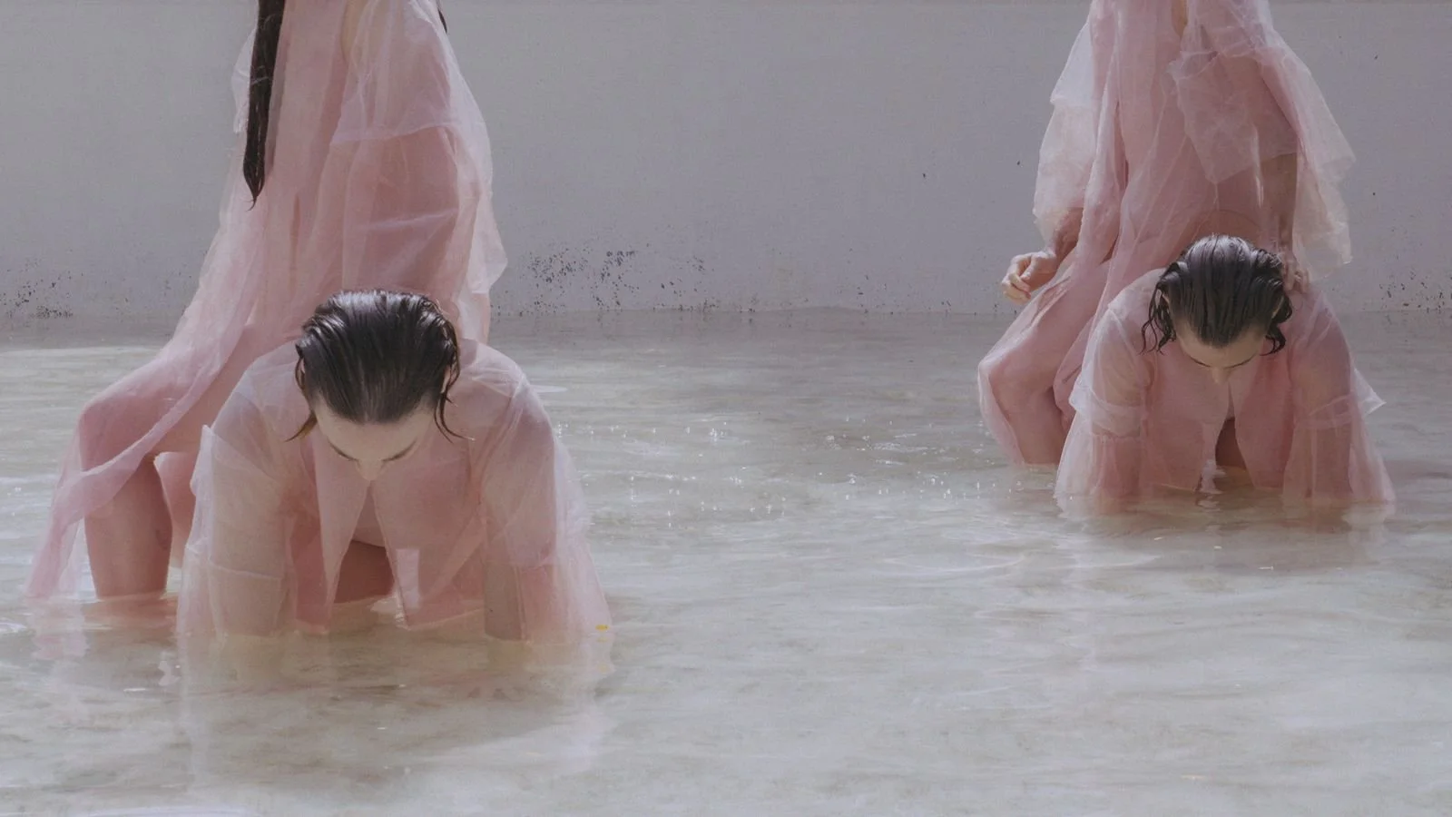 Four women in pink, sheer dresses with wet hair are crouching and kneeling in water indoors, setting a contemplative scene.