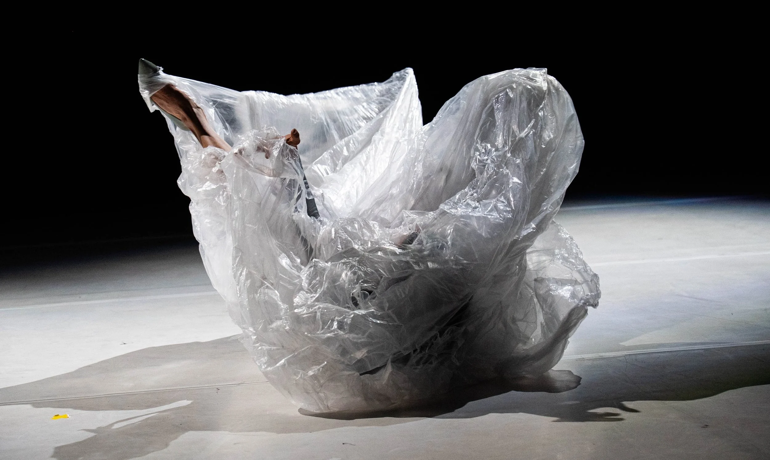 A person is sitting on the floor, wrapped in crumpled clear plastic, with only their arms and head visible. The background is black, and the floor is light-colored.