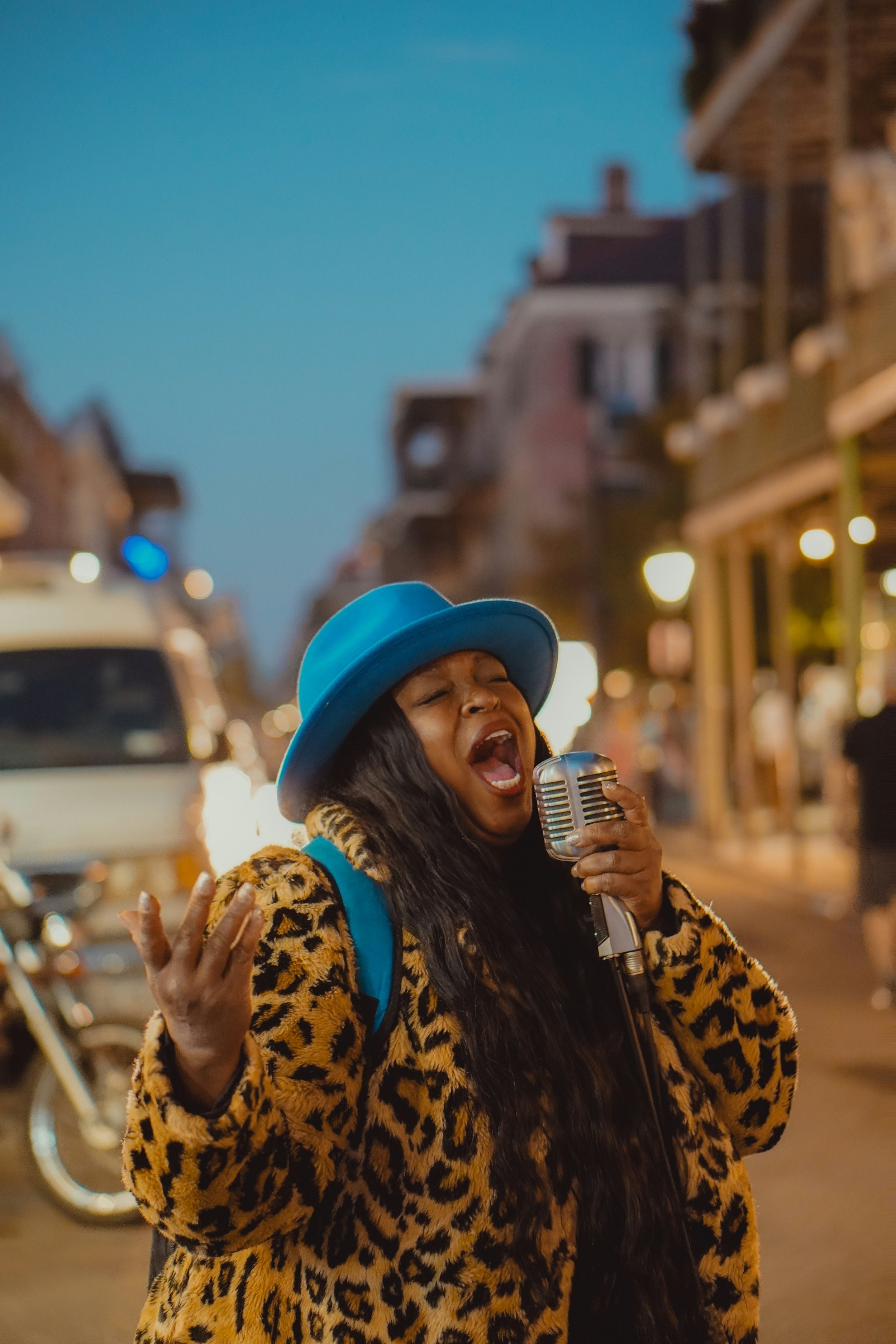 A musician singing into a vintage microphone on a city street at night, wearing a blue hat and a leopard print coat.