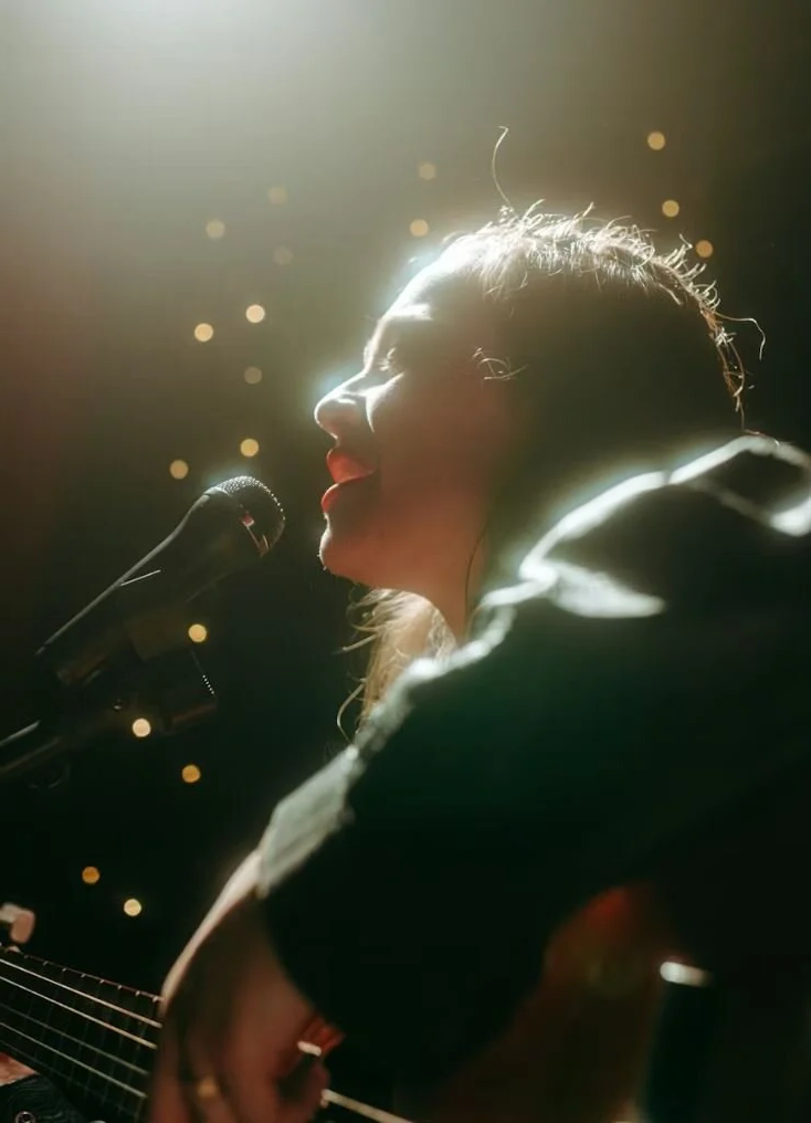 A woman singing into a microphone while playing guitar, illuminated by stage lighting with a blurred background.