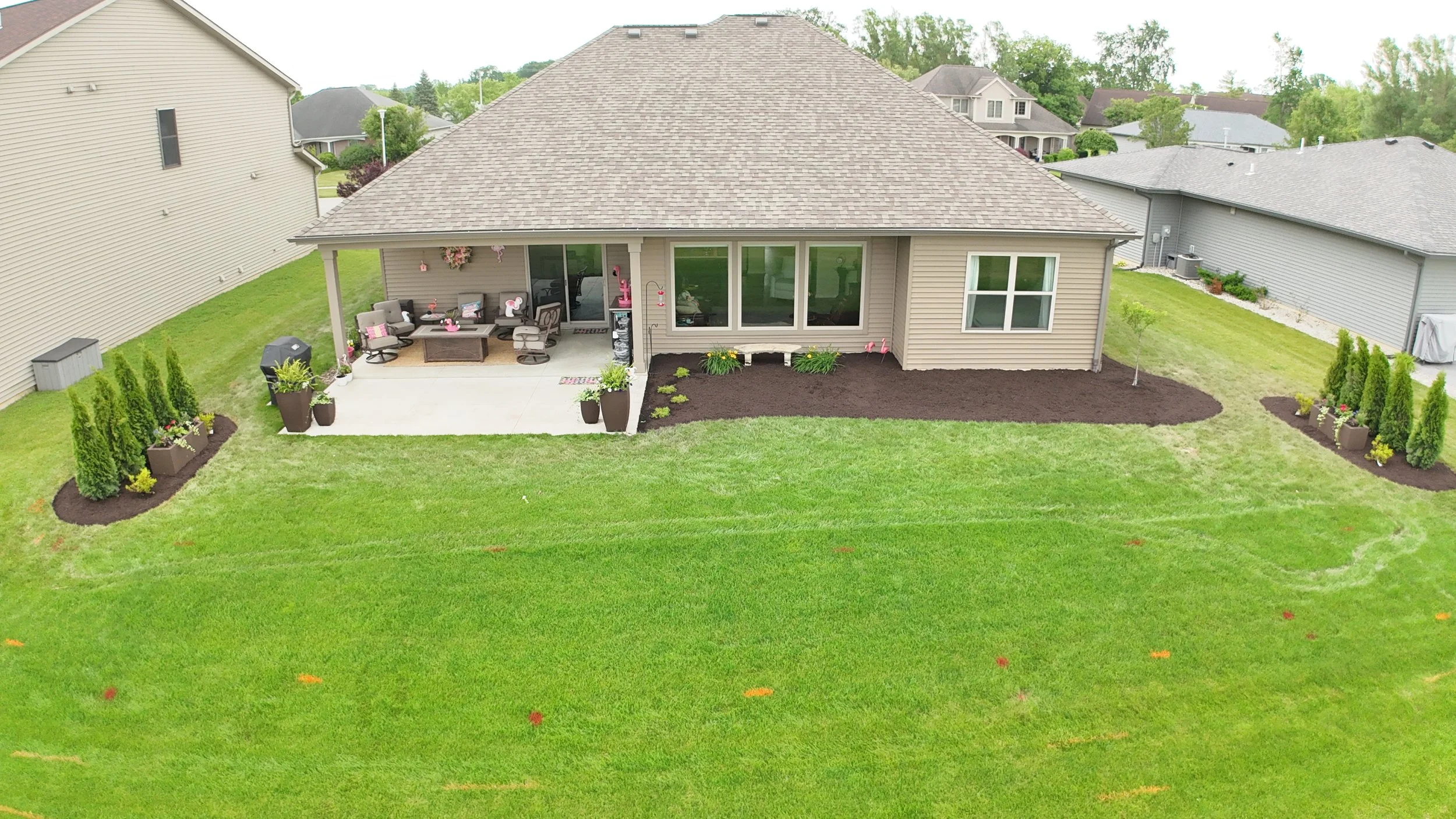 Backyard of a house with a patio, outdoor furniture, potted plants, and landscaped flower beds with mulch and small shrubs, surrounded by green lawn and neighboring houses.