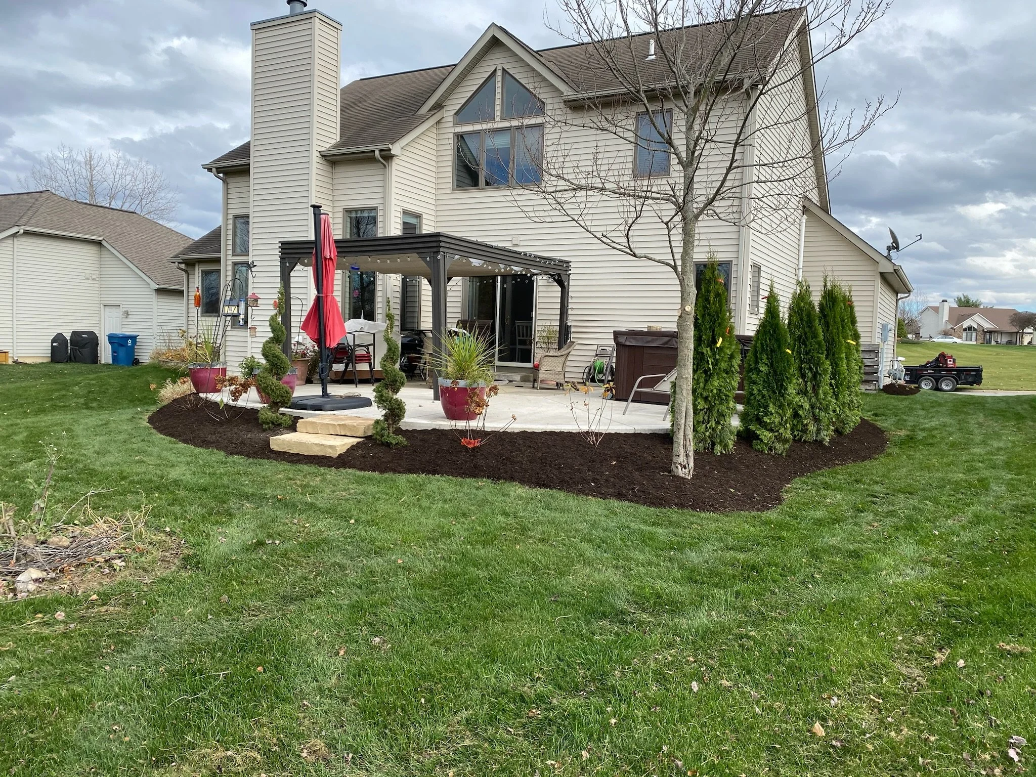 Backyard of a suburban house with a gray roof and beige siding, featuring a patio area with a pergola, red umbrellas, potted plants, a hot tub, and landscaping with trees and shrubs around the perimeter, set in a green lawn under a cloudy sky.