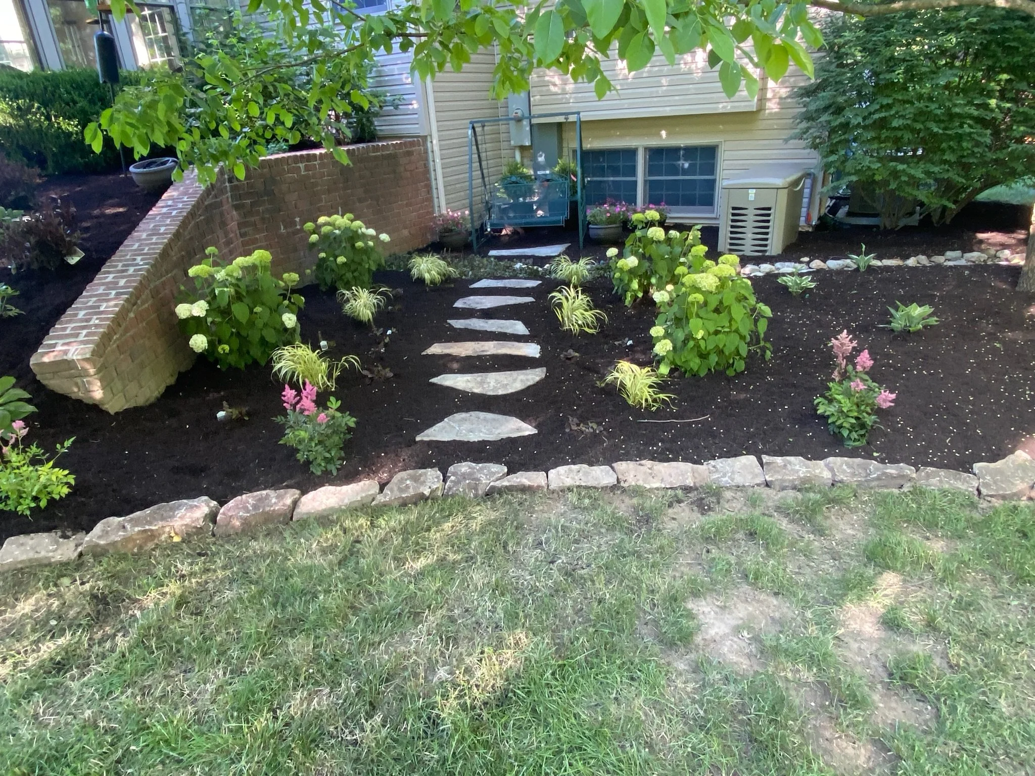 Landscaped garden with stone pathway, bordered by flowering plants, brick retaining wall, and shrubs, adjacent to a building.