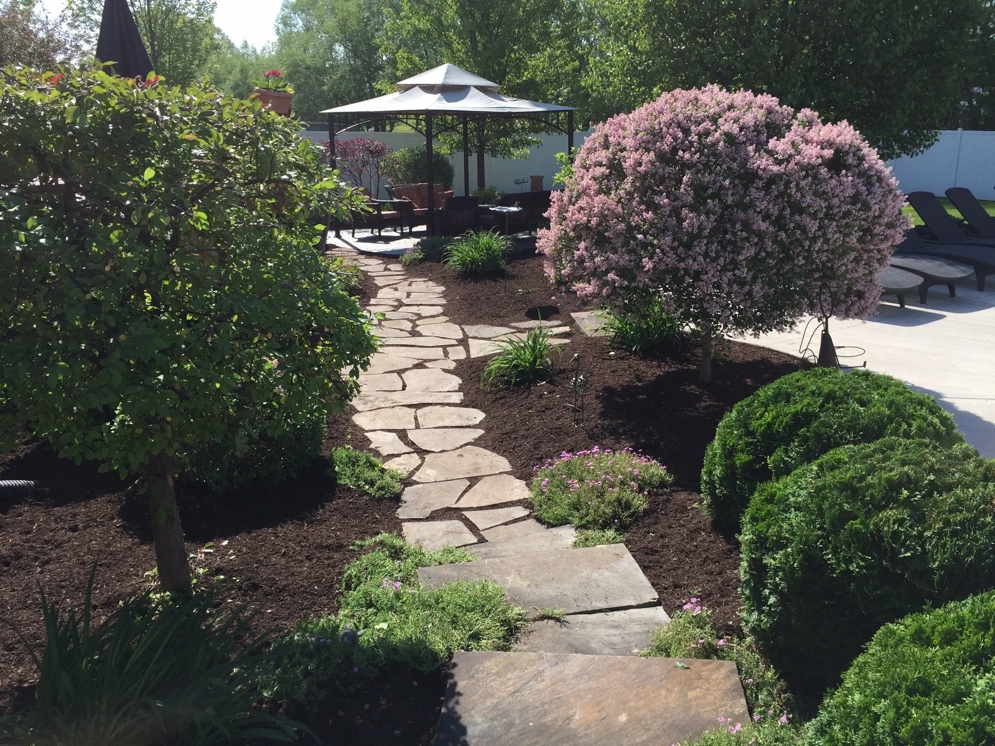 Garden pathway with stepping stones, surrounded by flowering shrubs and neatly trimmed bushes, leading to a seating area with patio umbrellas and lounge chairs.