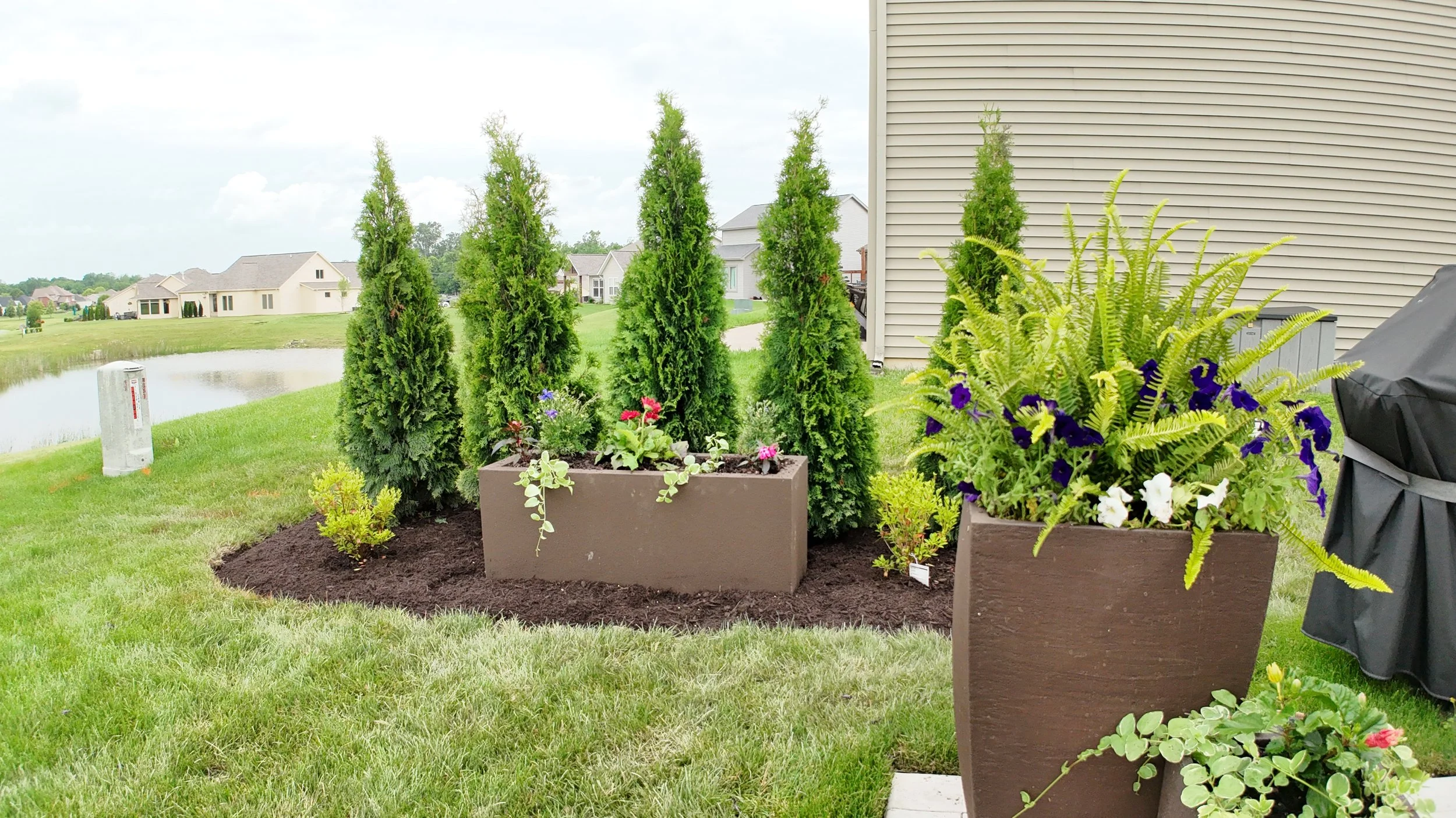 A well-maintained backyard garden with potted plants, green trees, and a view of houses and a pond in the background.