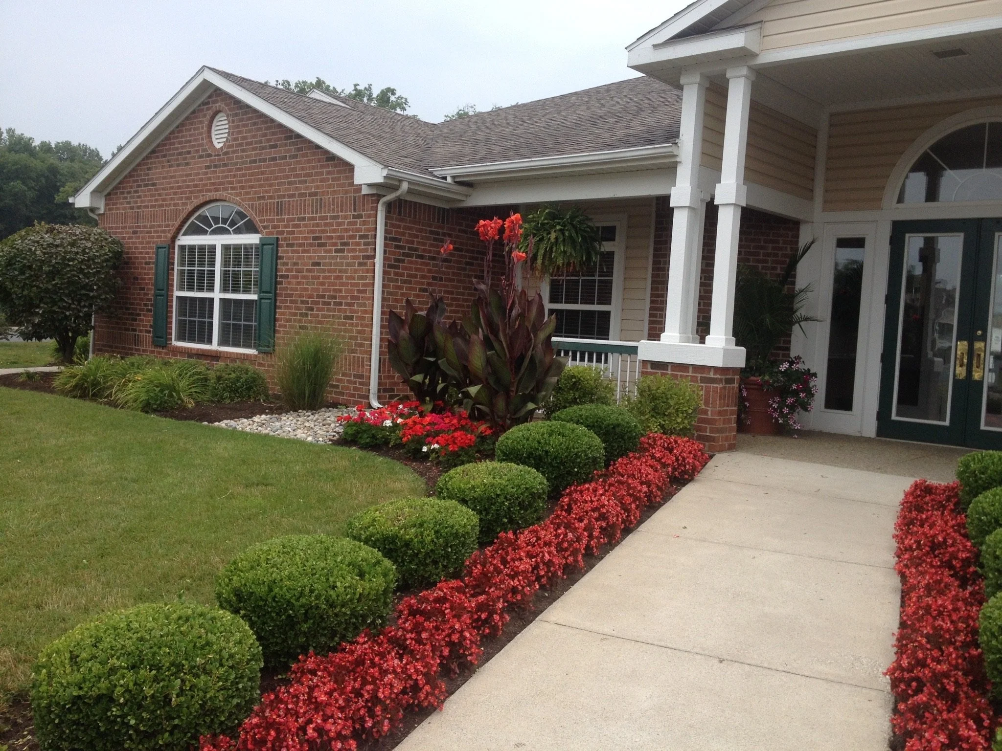 A brick house with a well-maintained garden, featuring a green lawn, neatly trimmed bushes, and red flowering plants alongside a concrete pathway leading to a porch with white pillars and dual doors.