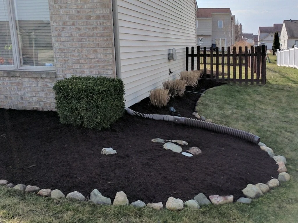 Landscape with mulched flower bed, shrubs, rock border, drainage pipe, and fence along house.