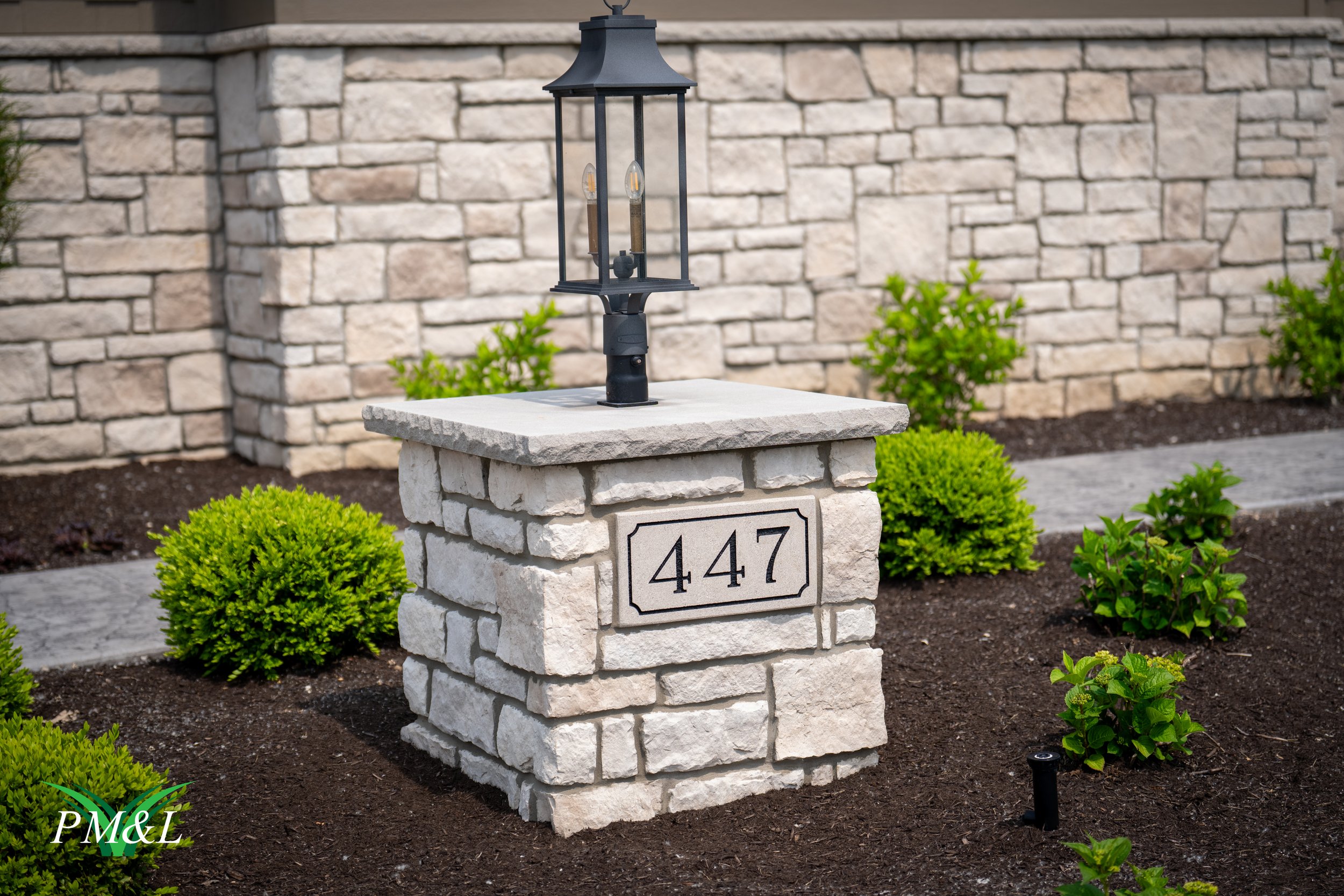 A stone mailbox with the house number 447 engraved on the front, topped with a black lantern-style lamp, set in a landscaped yard with green bushes and mulch, with a stone wall in the background.