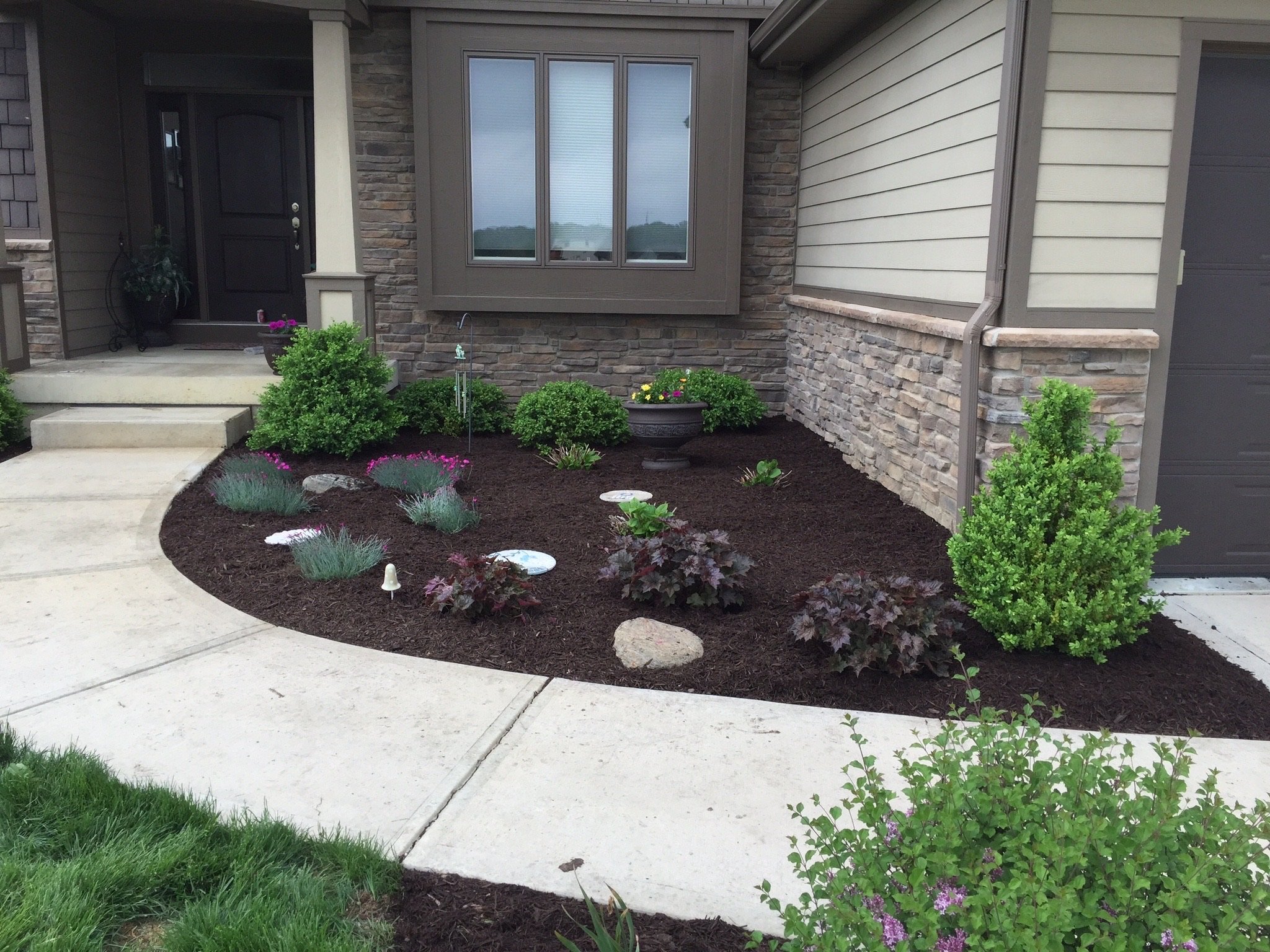 Front yard landscaping with shrubs, flowers, mulch, and a stone pathway near a house entrance.