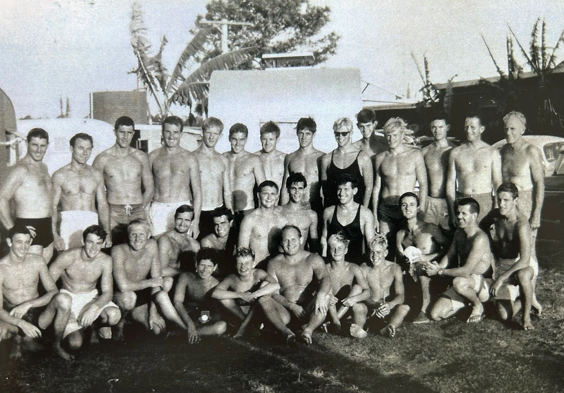 Group of young men and women in swimsuits at a beach, some sitting and some standing, with boats and palm trees in the background, in a black-and-white photo.