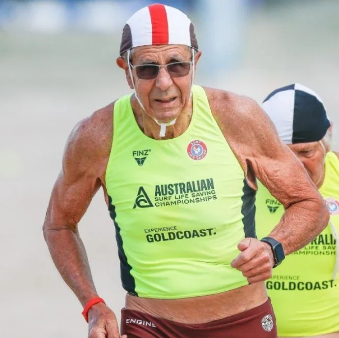 An elderly male athlete participating in the Australian Surf Life Saving Championships, wearing a neon yellow tank top, maroon shorts, sunglasses, and a red, white, and black swim cap, running on the beach.