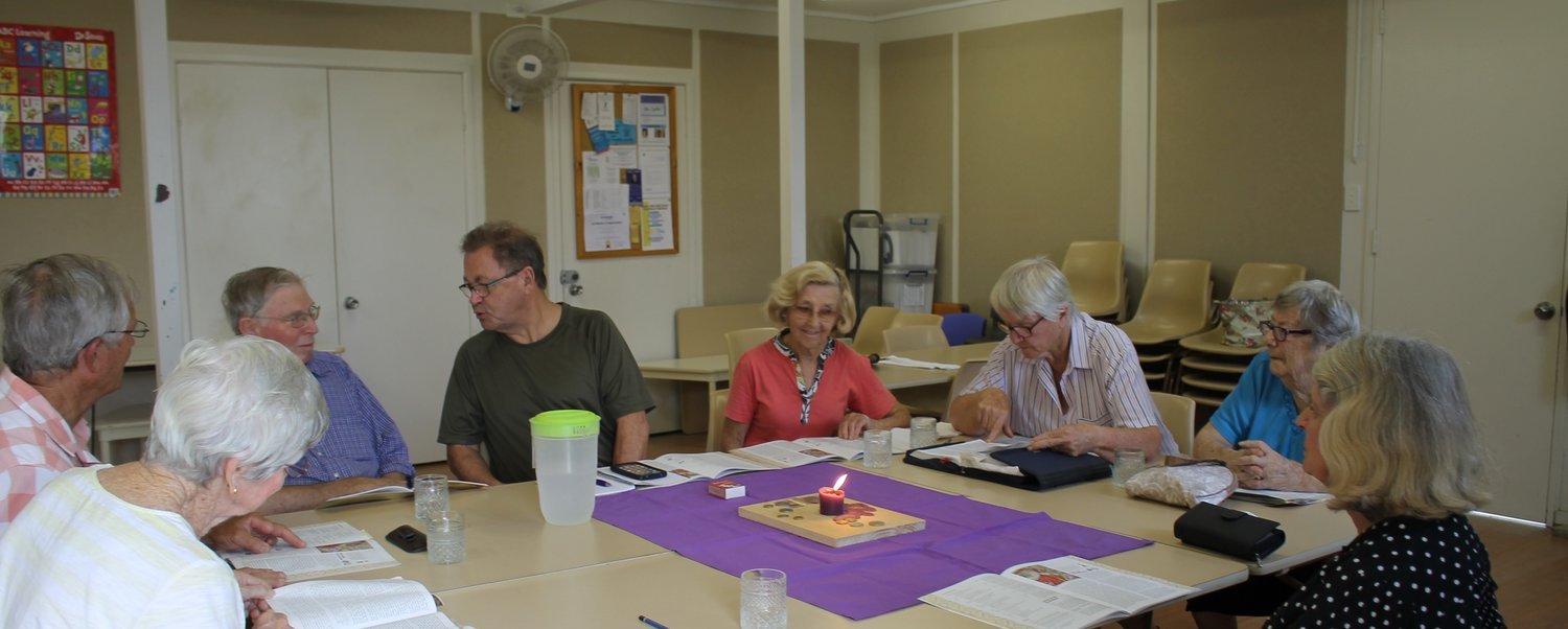 Group of elderly people sitting around a table in a room, reading books or materials, with a lit candle on the table, in a community setting.
