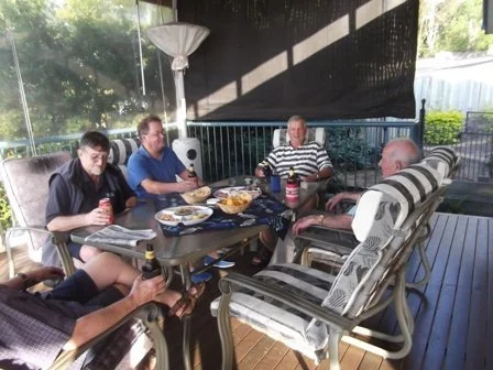 Five people sitting around a table enjoying food and drinks on a covered patio with outdoor furniture and trees visible in the background.