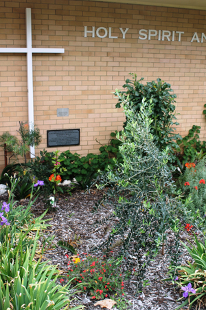 Church garden with a large cross and the words 'HOLY SPIRIT AND' on a brick wall, surrounded by various plants and flowers.