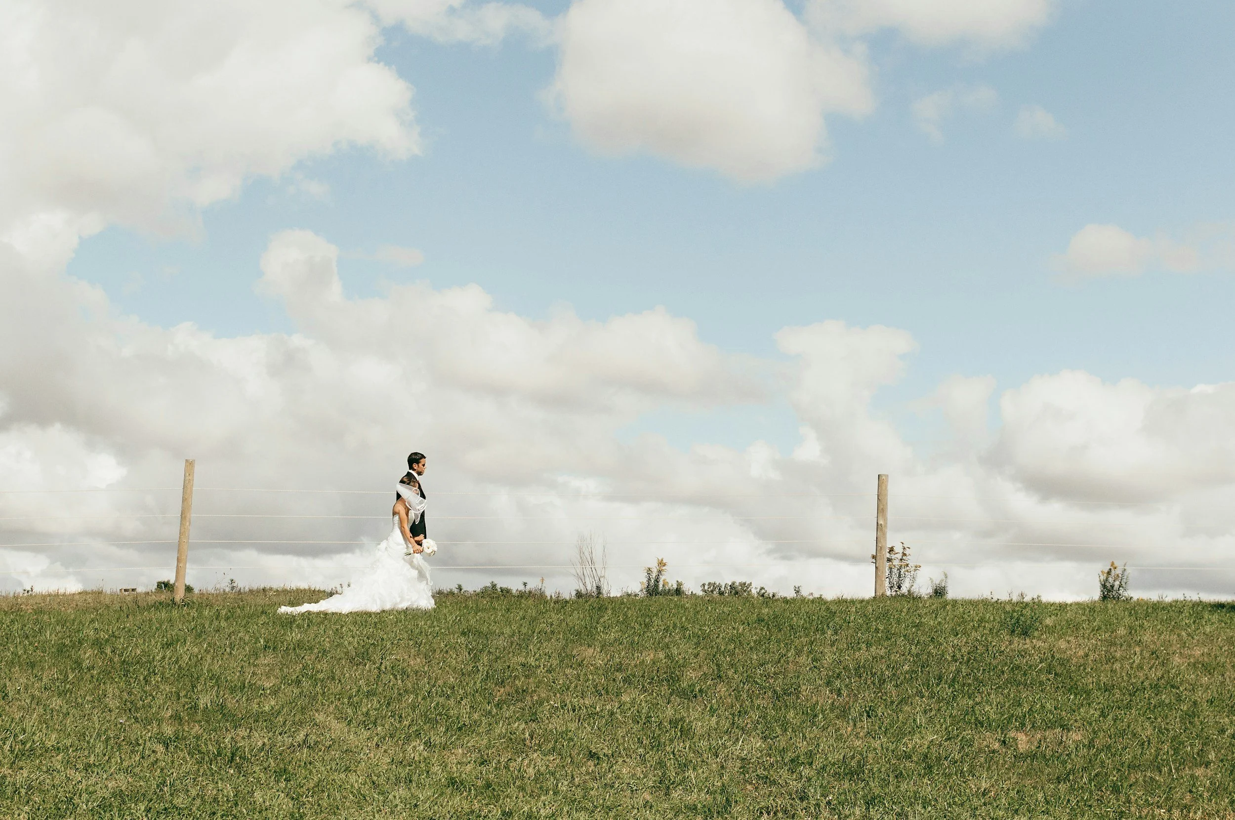 A bride and groom walking together on a grassy field with a fence and cloudy sky in the background.