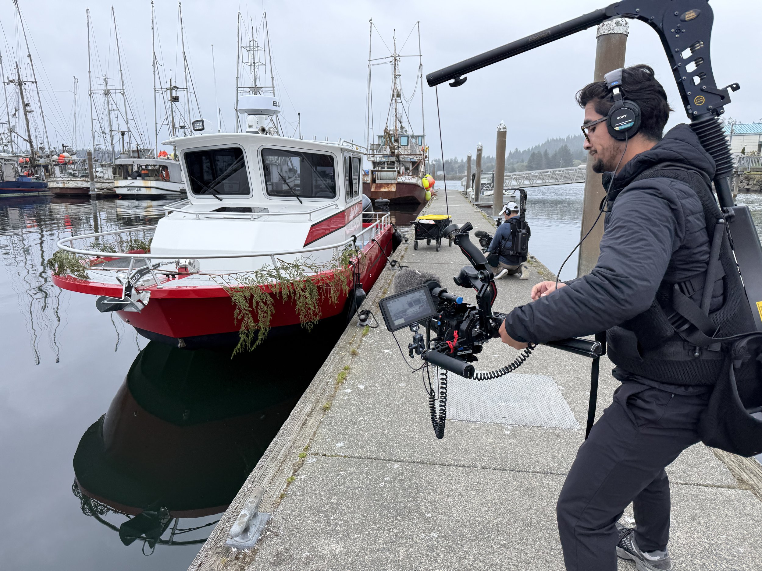 A man with dark hair, glasses, and a beard wearing a black jacket, headphones, and carrying camera equipment stands on a dock next to a boat, with several other boats and sailboats docked in the background on a cloudy day.