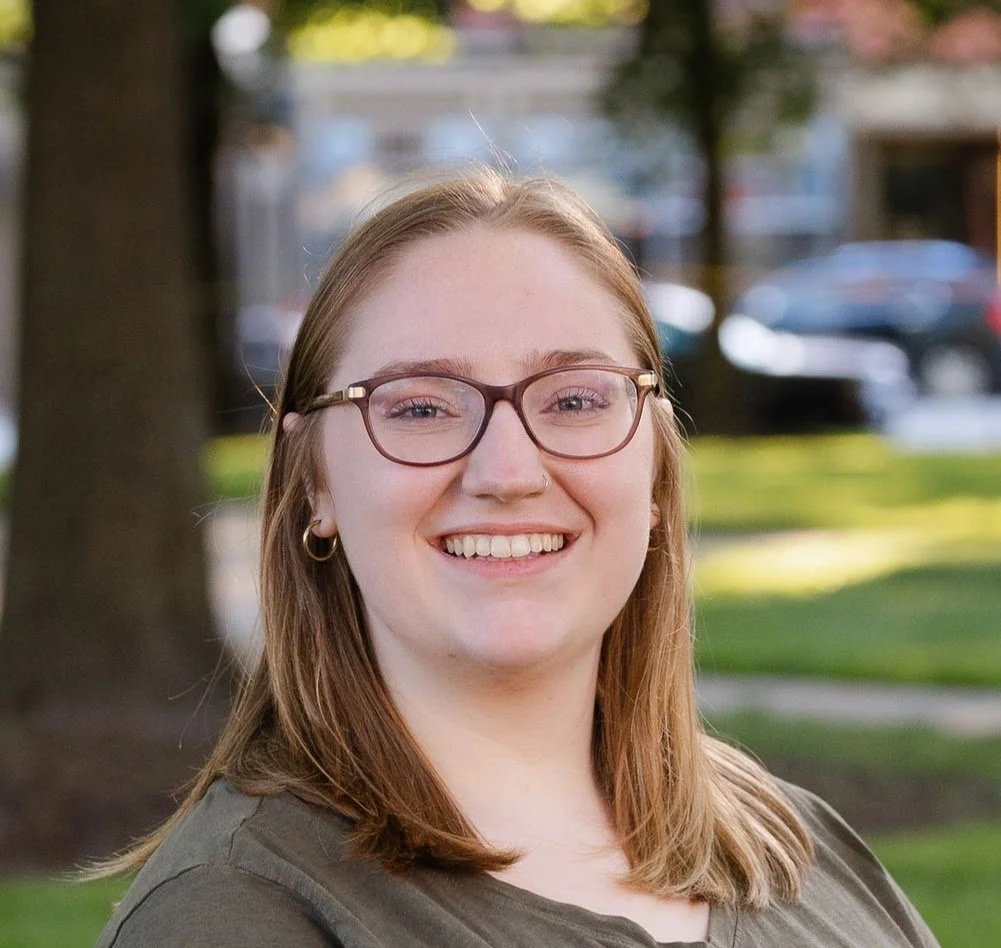 Smiling woman with light brown hair, glasses, gold hoop earrings, and a nose ring, standing outdoors with trees and a parking lot in the background.