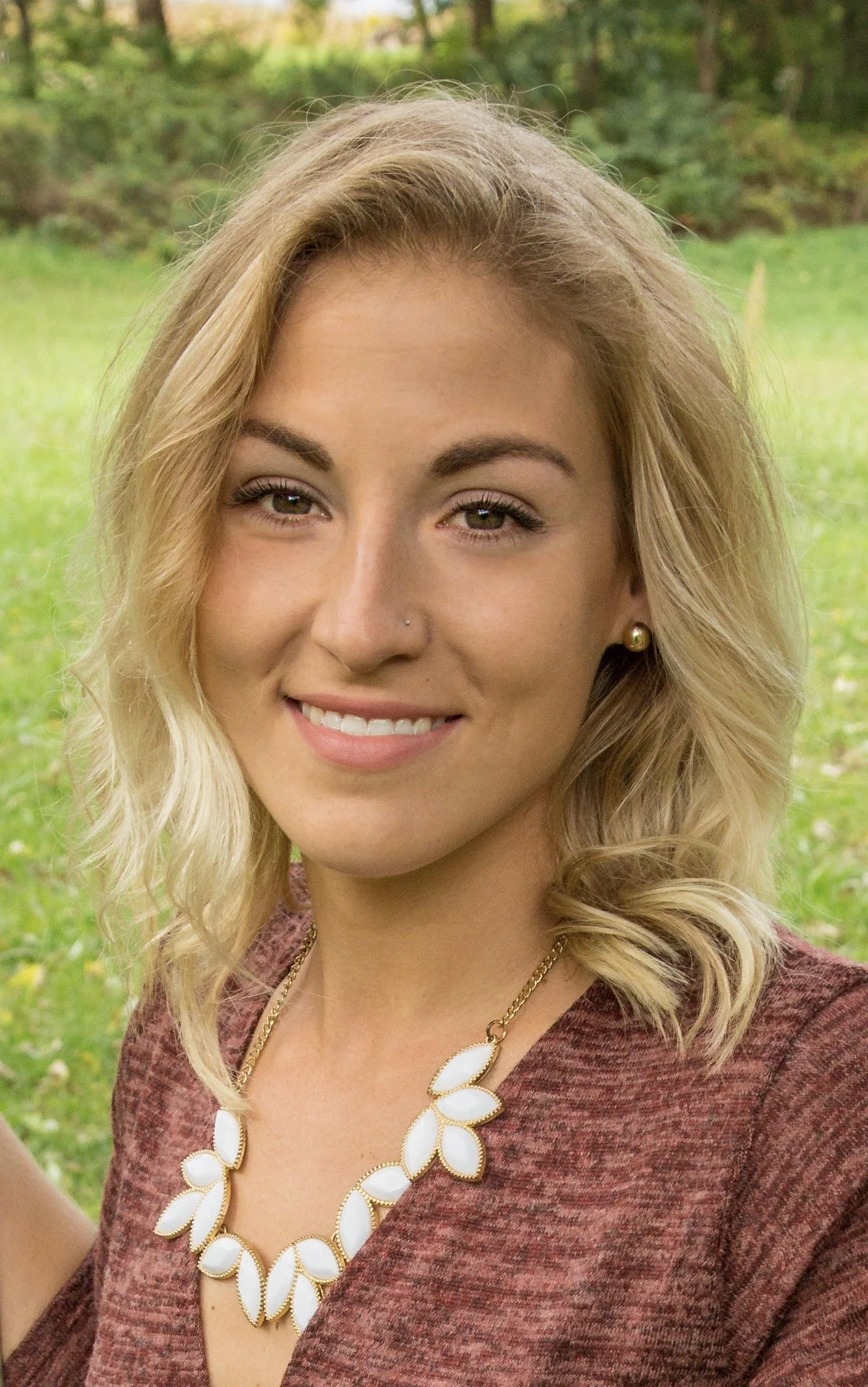 A smiling young woman with blonde, wavy hair, wearing a maroon top, a white leaf-shaped necklace, and pearl earrings, outdoors with greenery in the background.