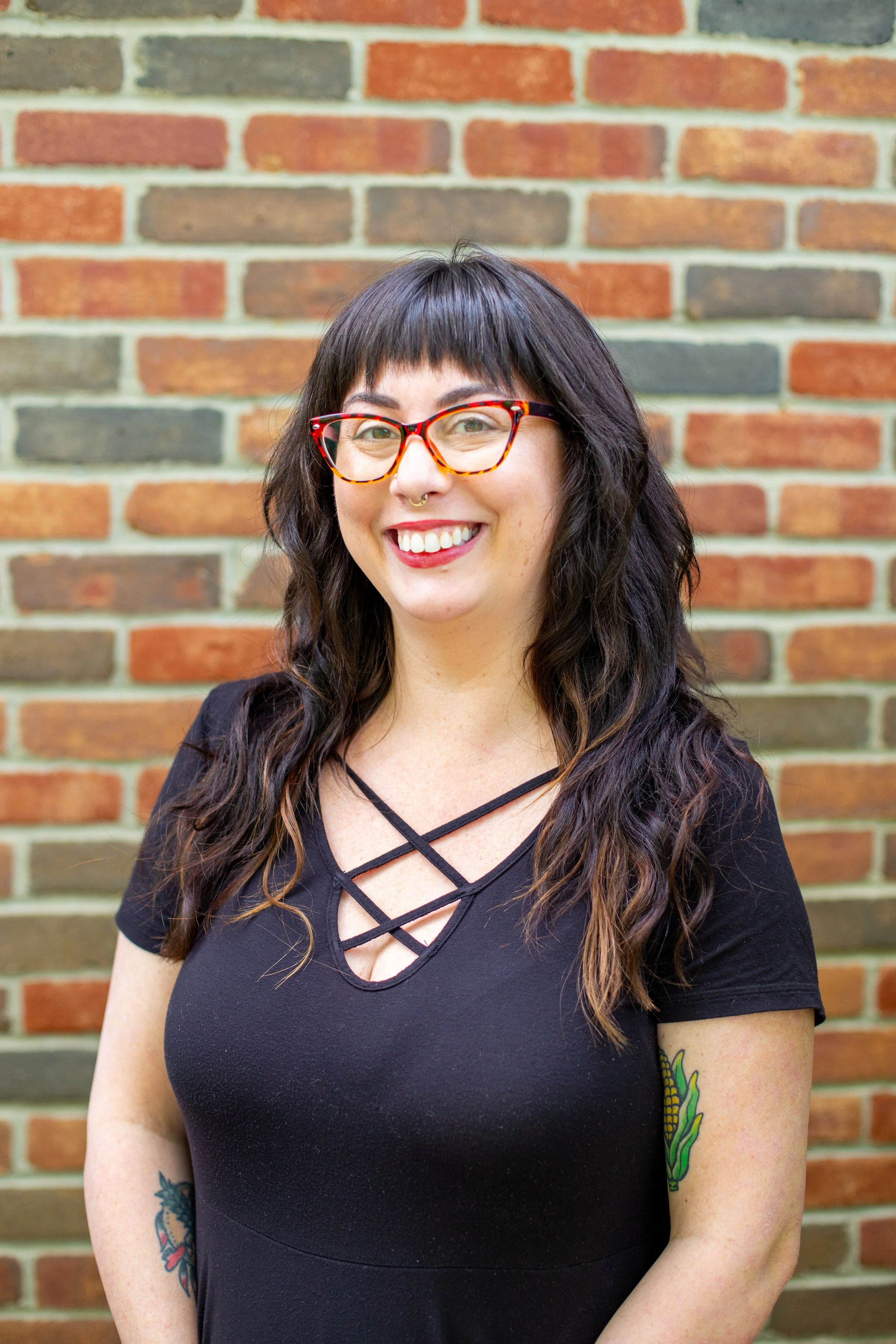 A woman with dark wavy hair, red glasses, and tattoos on her arms, smiling in front of a brick wall.