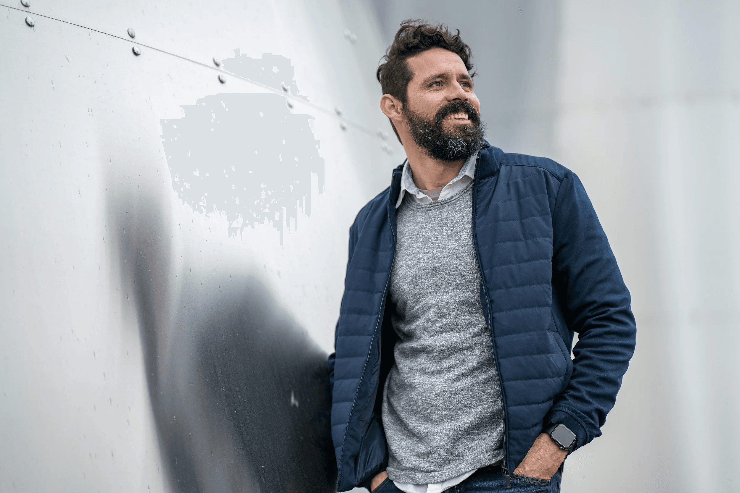 A man with dark curly hair and a beard smiling while leaning against a metallic wall, wearing a navy blue jacket, gray sweater, white shirt, and a smartwatch.