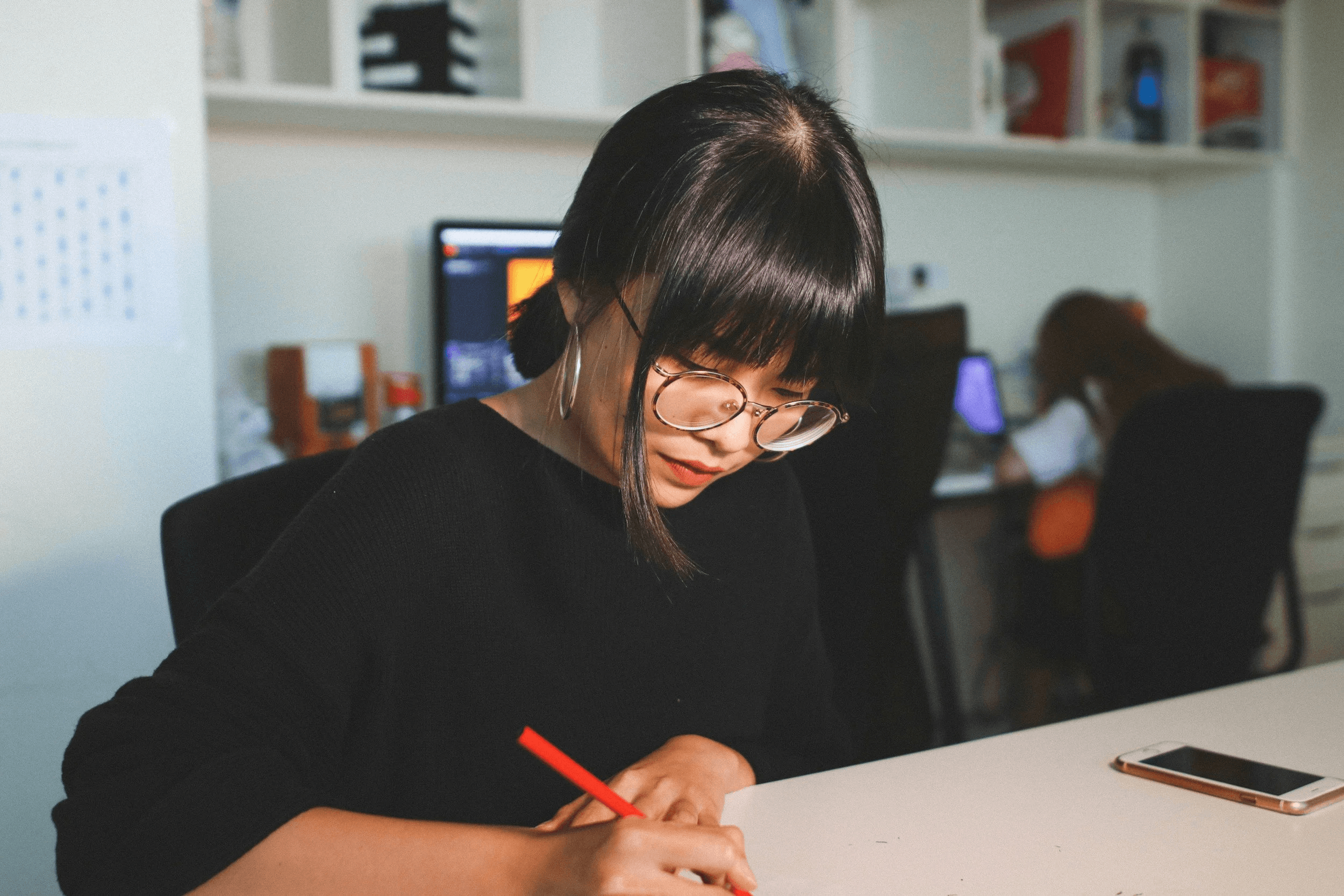 A woman wearing glasses and hoop earrings sits at a desk writing with a red pen, with a smartphone on the desk and a computer monitor in the background.